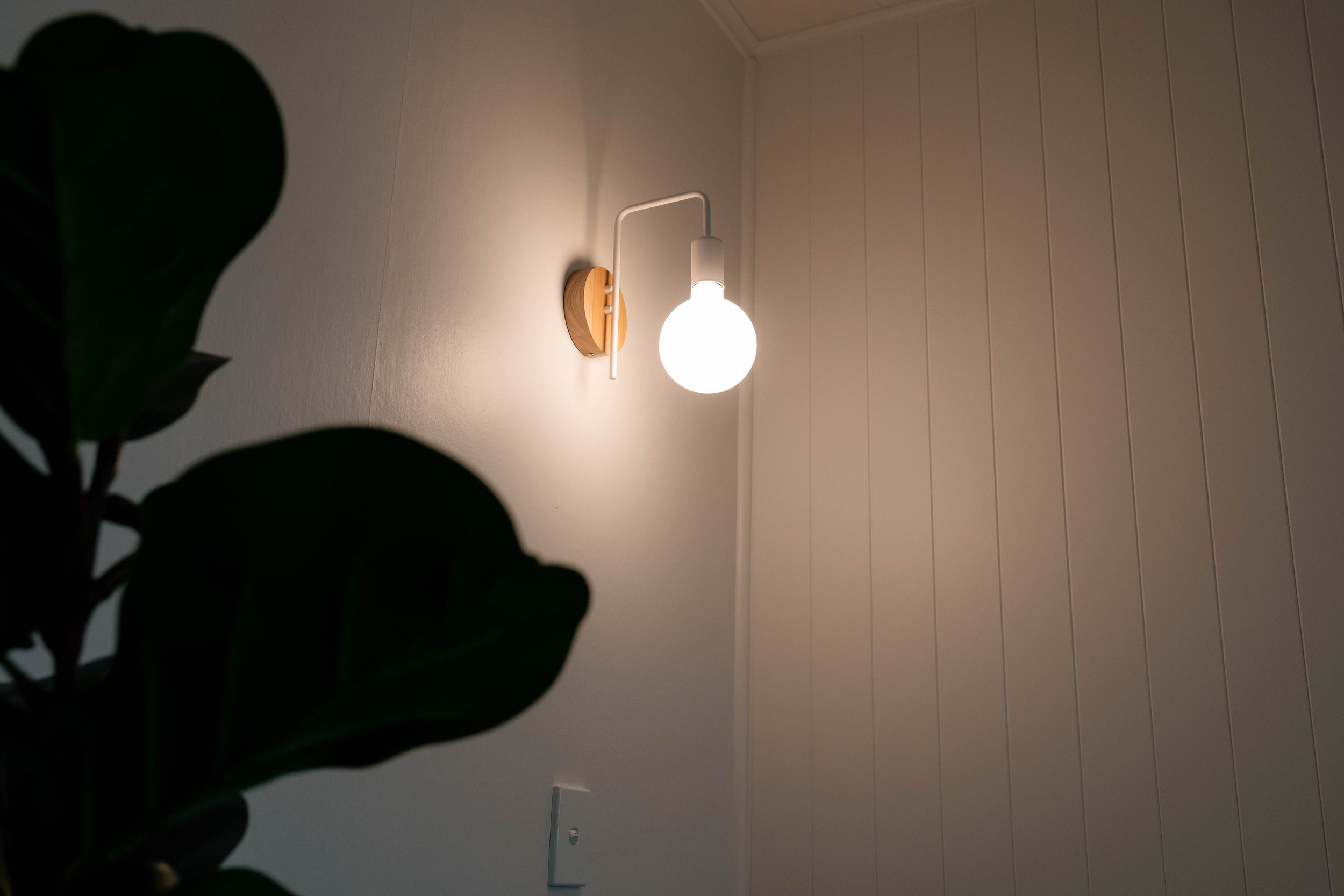 A wall-mounted white lamp with a wooden base illuminated against a beige paneled wall, with dark green leaves partially obscuring the bottom left corner.