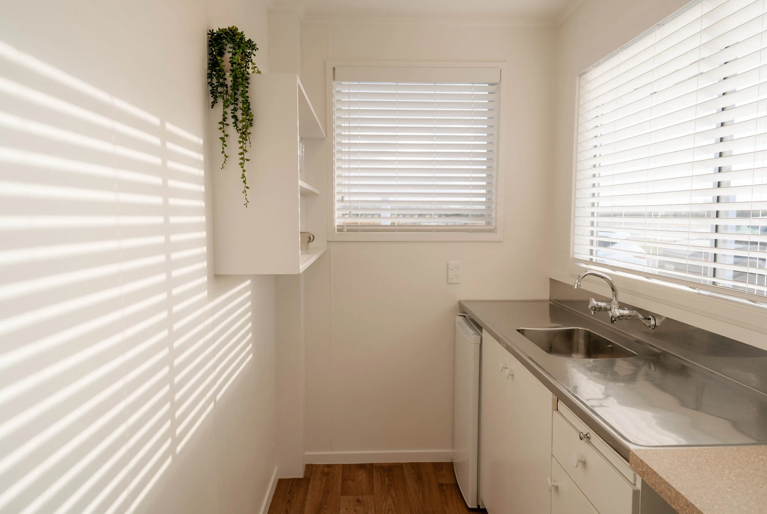 A small kitchen with white cabinets, a stainless steel sink, and large windows with blinds casting striped shadows on the wall.