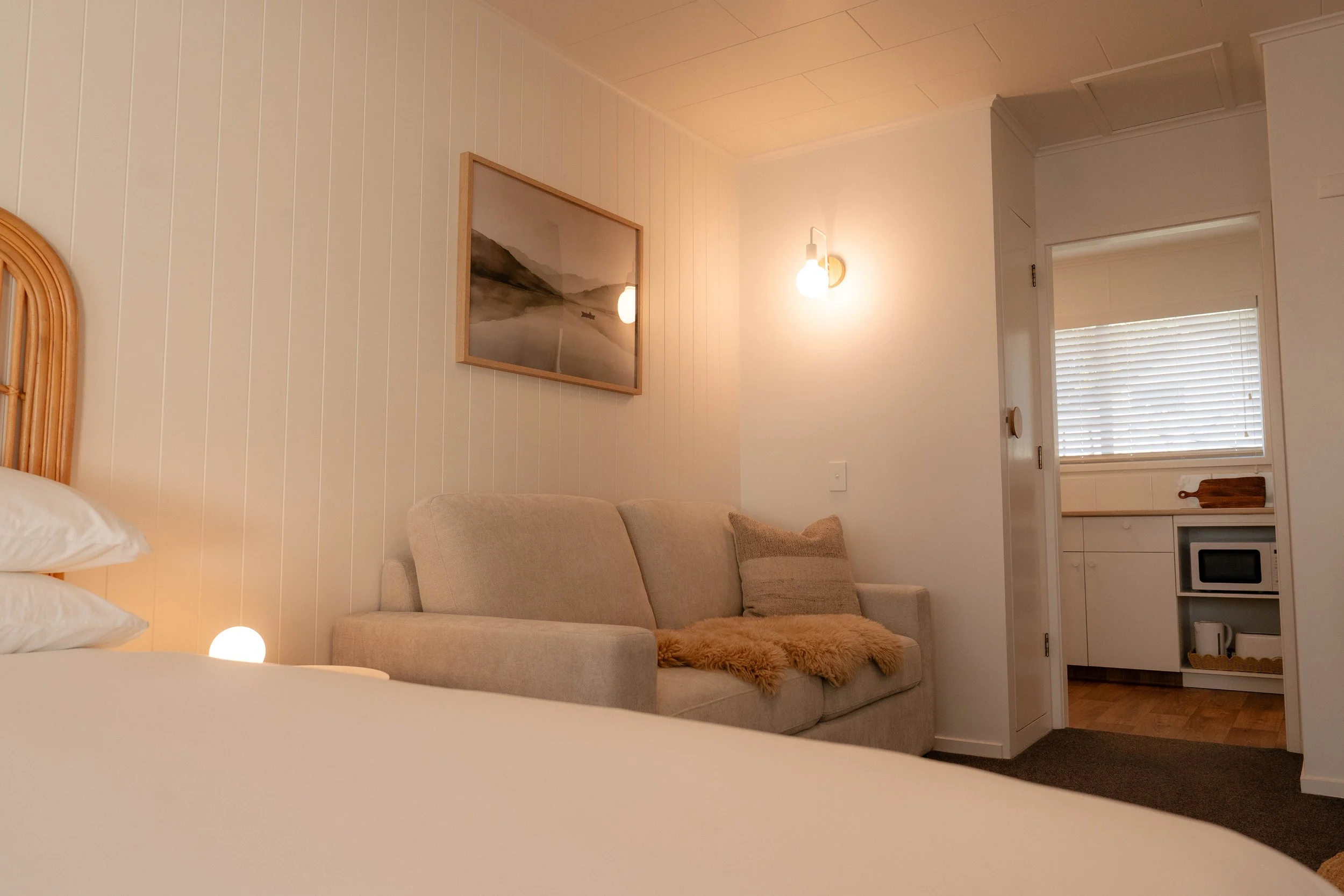 A cozy living room with a beige sofa, a furry throw blanket, wall-mounted lights, framed artwork, and a small kitchen area with blinds in the background.