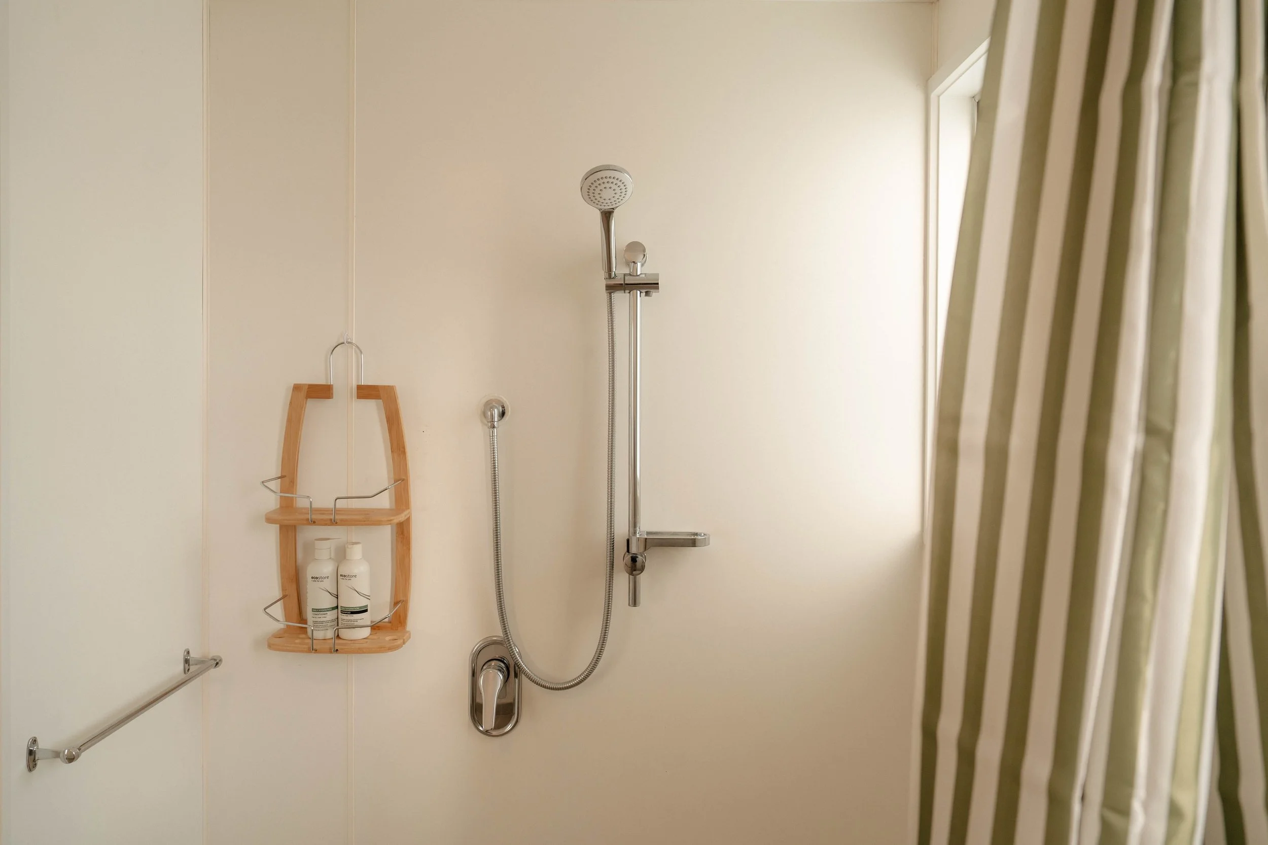 Shower area with handheld showerhead, soap holders, and bottles on a wooden shelf, partial view of a shower curtain to the right.