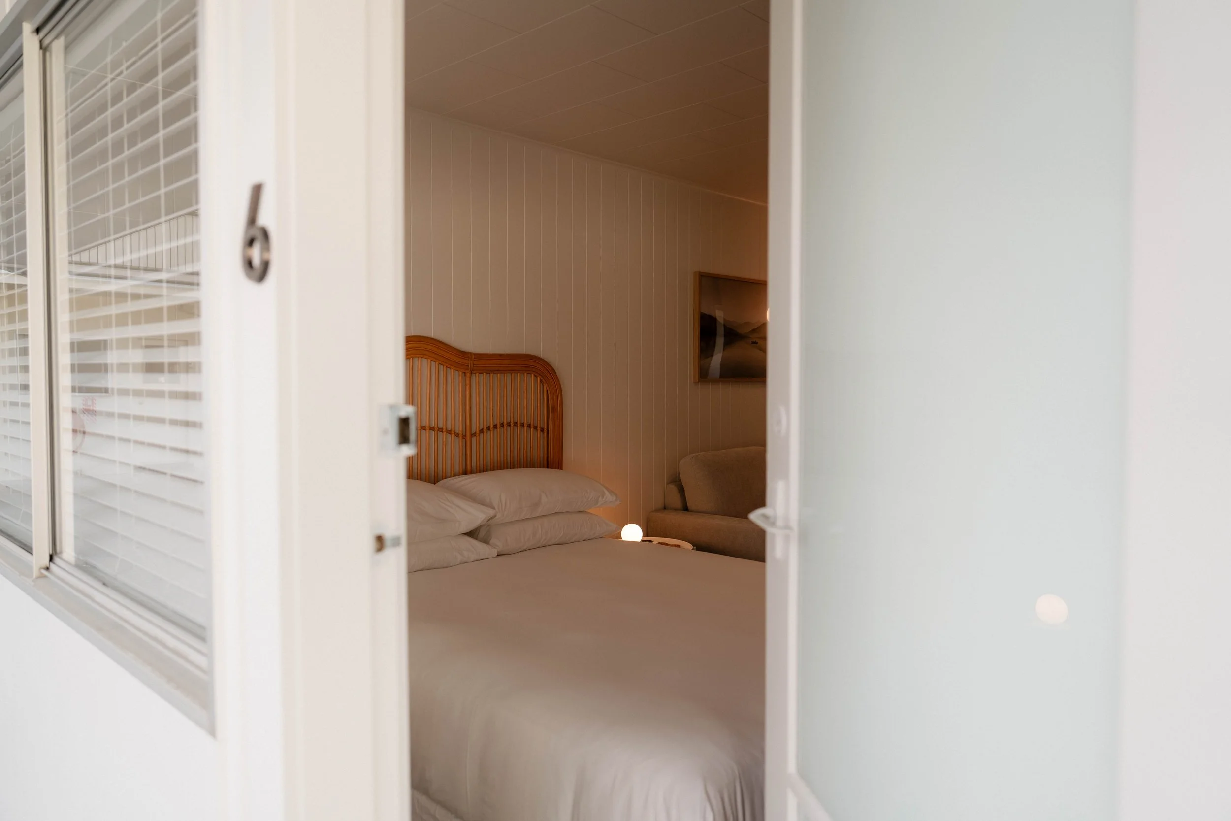 Bedroom viewed through partially open window with white blinds, showing a bed with white linens, a wooden headboard, a wall-mounted landscape photograph, and a small round lamp on a nightstand.