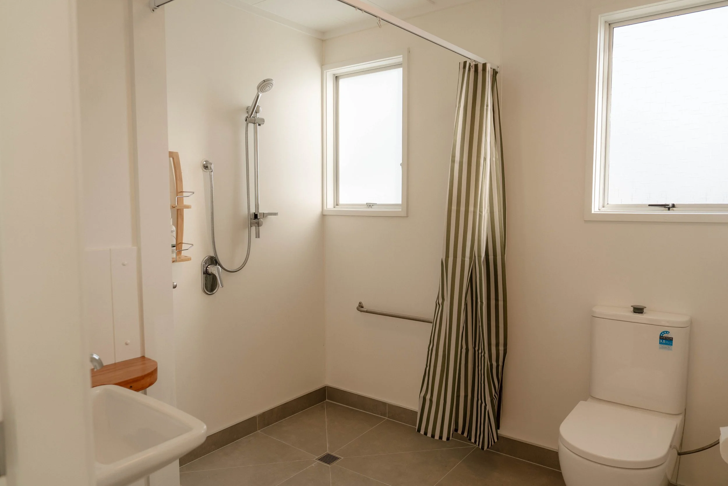 Bathroom with a shower, two windows, a toilet, and a small sink. The shower has a striped curtain, a showerhead, and a wooden shelf. The floor has gray tiles and a floor drain.