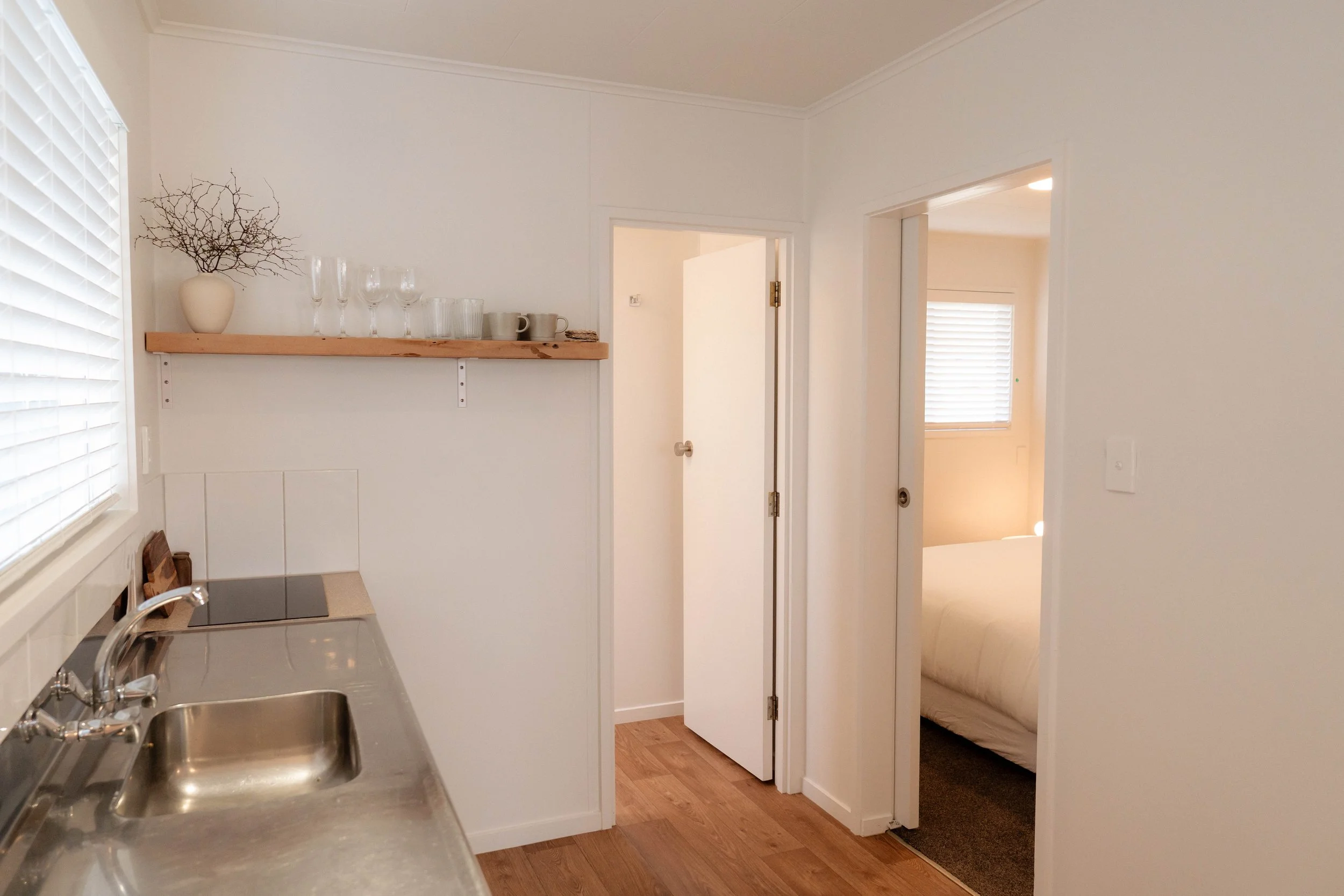 A view of a small kitchen with a sink, a window with blinds, and a wooden shelf with glassware and a vase with twigs. There is a door leading to a bedroom with a window and blinds.