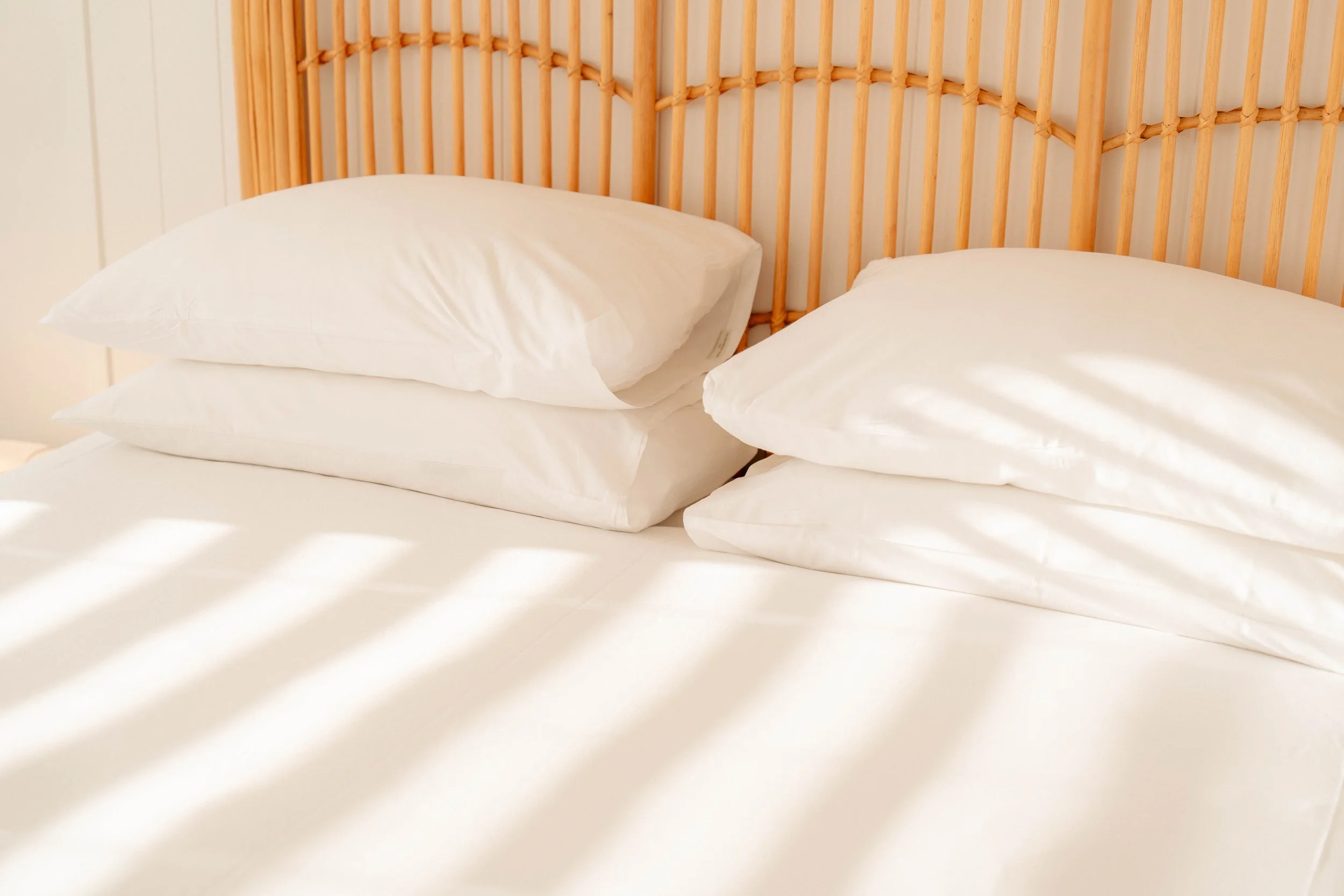 Close-up of a bed with white sheets and pillows, wooden headboard, bright natural light casting shadows on the bed.