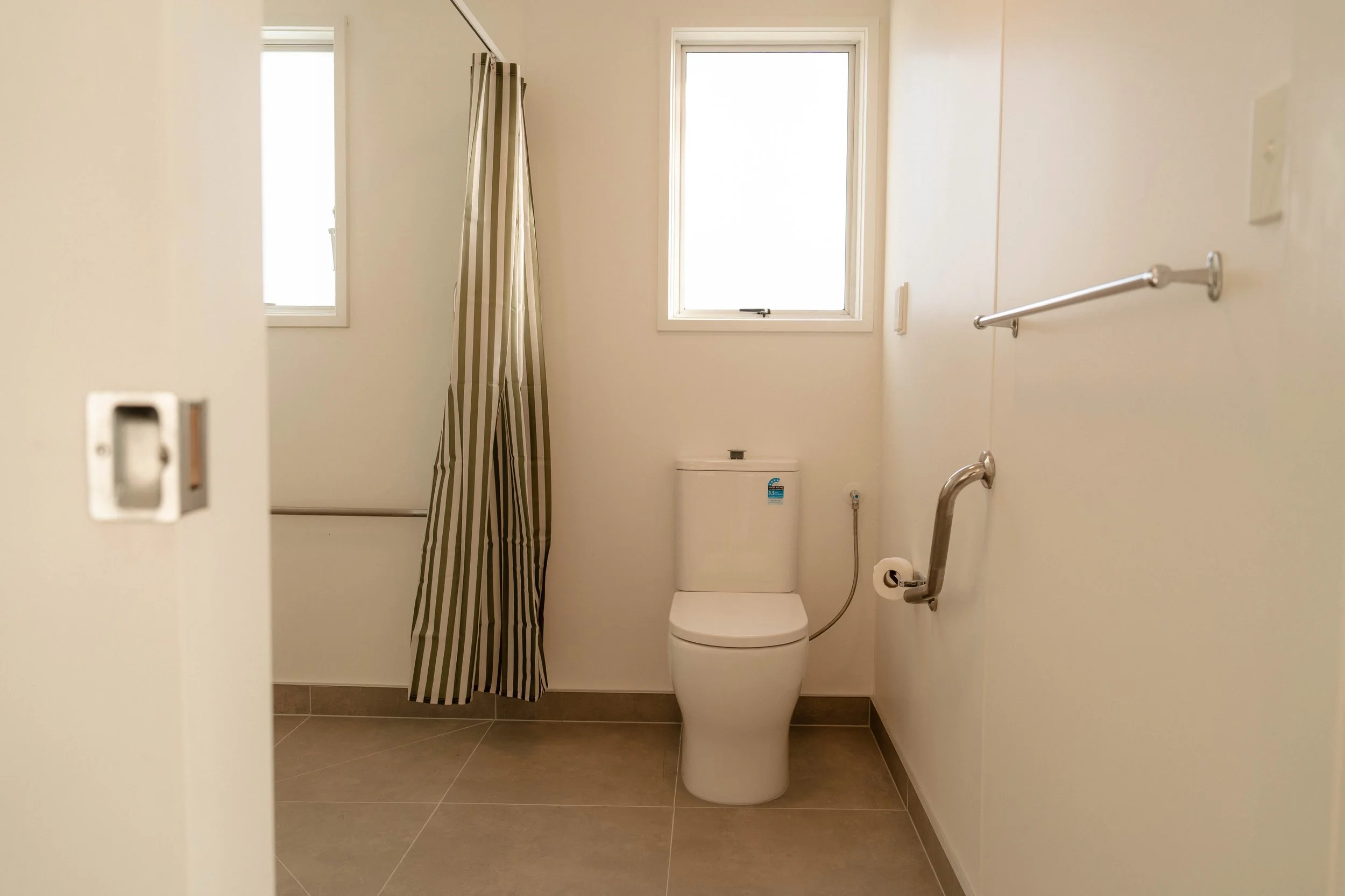 Small bathroom with a toilet, beige tile floor, cream-colored walls, two small windows, one with a striped shower curtain, grab bars on the wall, and a partial view of a doorframe.