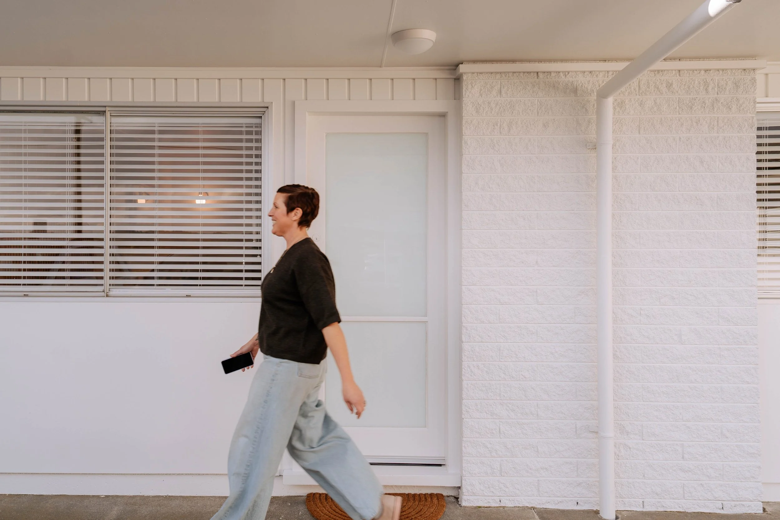 A woman with short brown hair wearing a black top and light blue jeans walking past a white brick wall and a window with closed blinds.