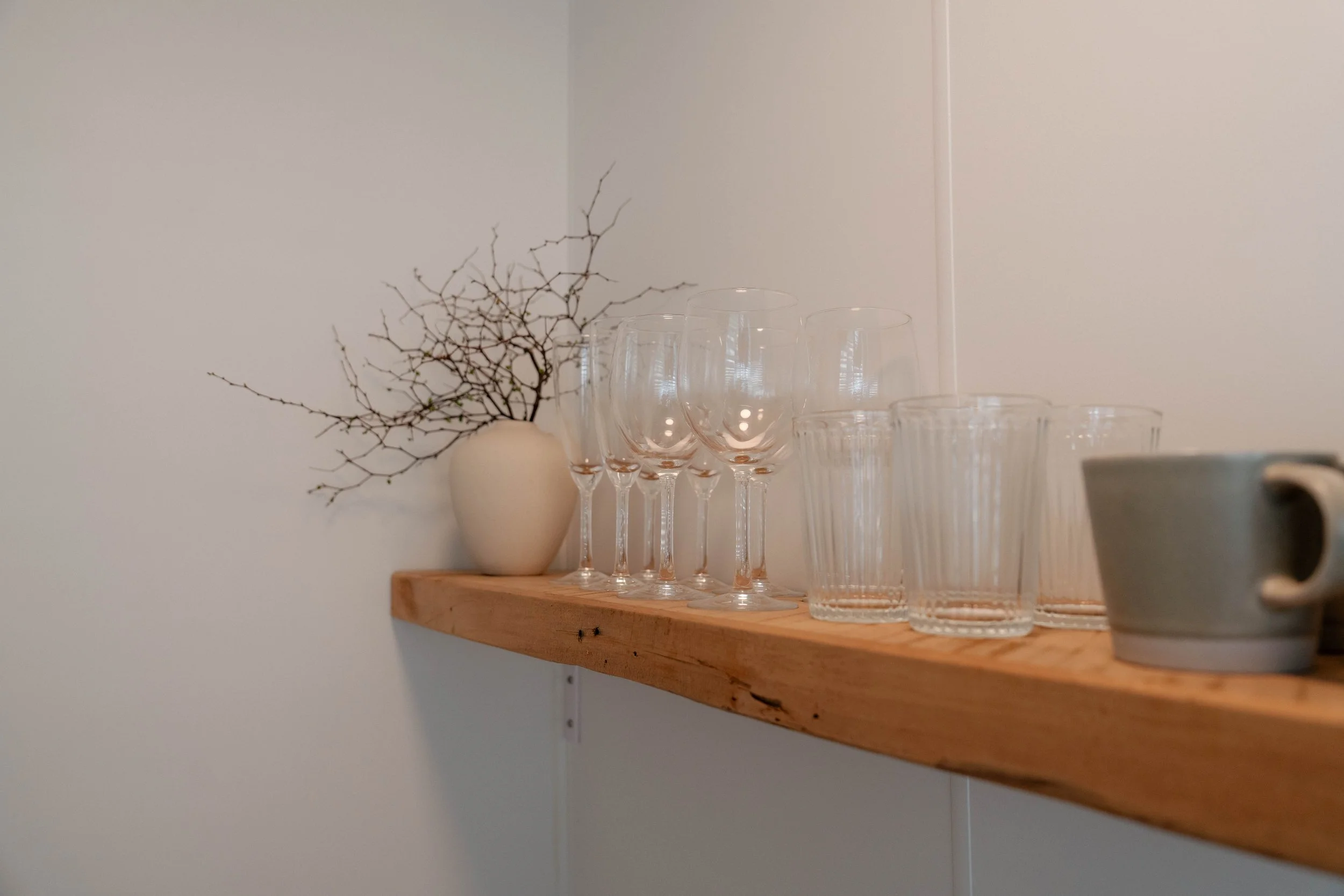 A wooden shelf with a white vase holding a dried branch, several empty wine glasses, and clear drinking glasses against a plain wall.