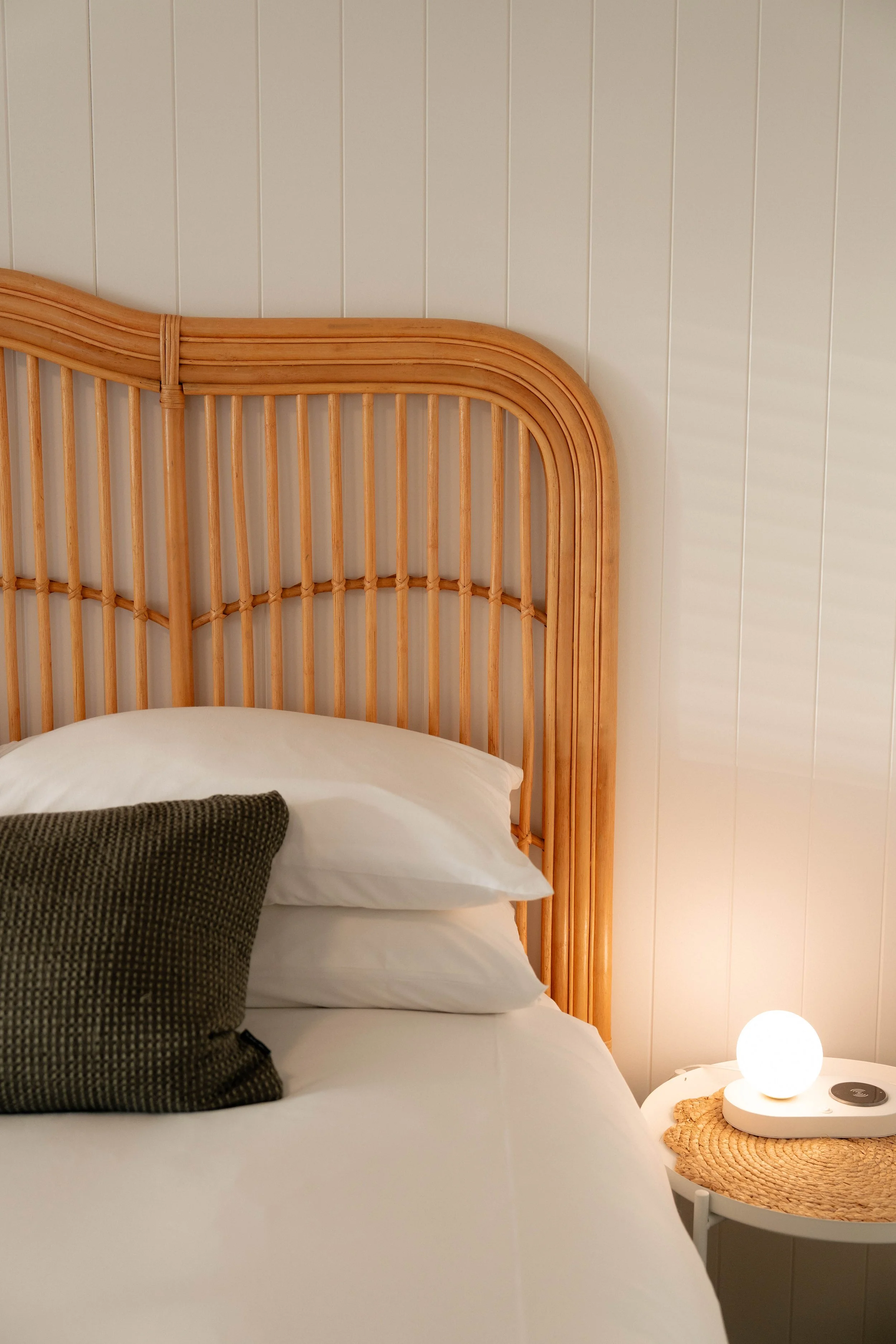 A bedroom with a carved wooden headboard, white pillows, a black-patterned throw pillow, a small white side table, a woven placemat, a spherical lamp, and a lamp on the side table. The wall is covered with light-colored vertical wood paneling.