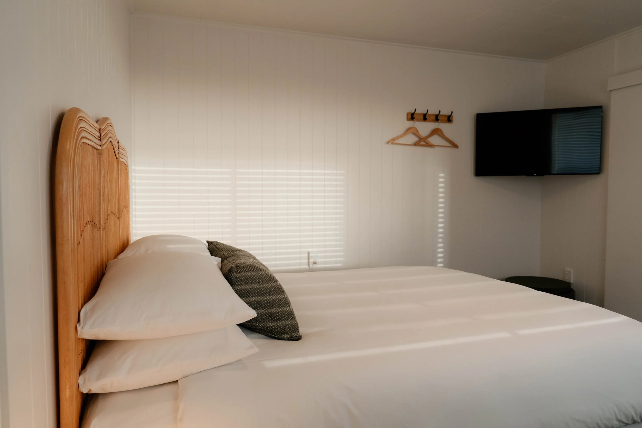 A bedroom with a wooden headboard, two pillows, a throw pillow, and a white bed sheet. There are plantation blinds on the window, a wall-mounted flat-screen TV, and a small coat rack with hangers.