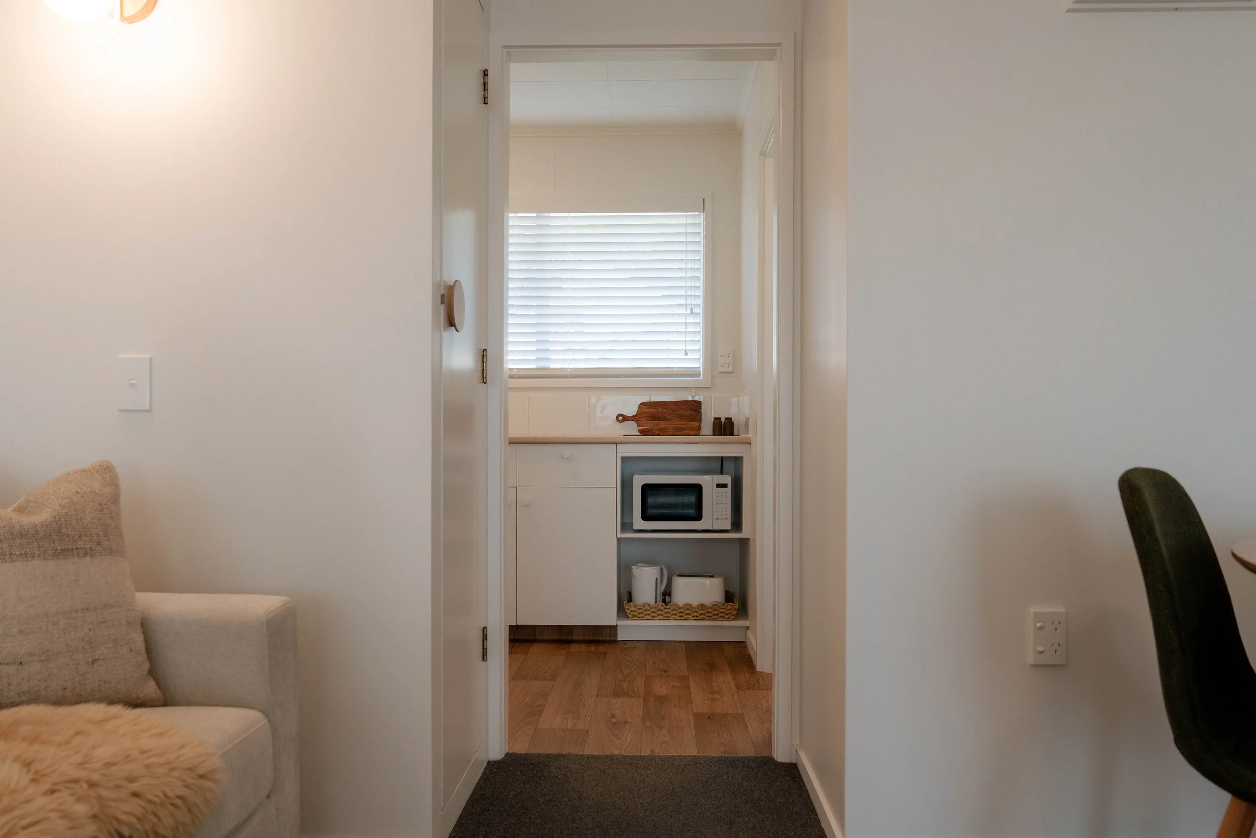 Interior view of a small kitchen with a microwave, a wooden cutting board, and window blinds visible through an open doorway from a living room with a beige sofa and a fuzzy blanket.