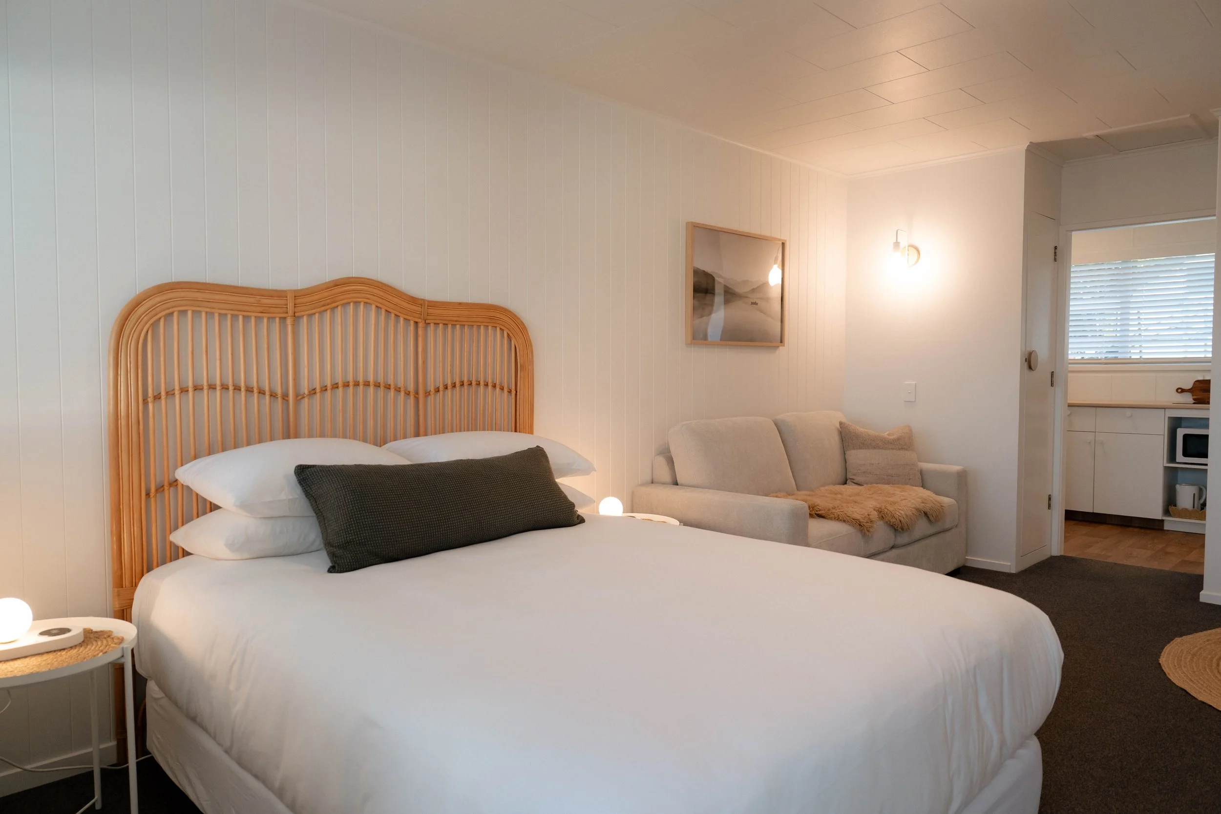 A cozy bedroom with a wicker headboard, white bedding, black pillows, a beige sofa with pillows and a fur throw, a framed landscape painting, a wall-mounted light, and a window with blinds.