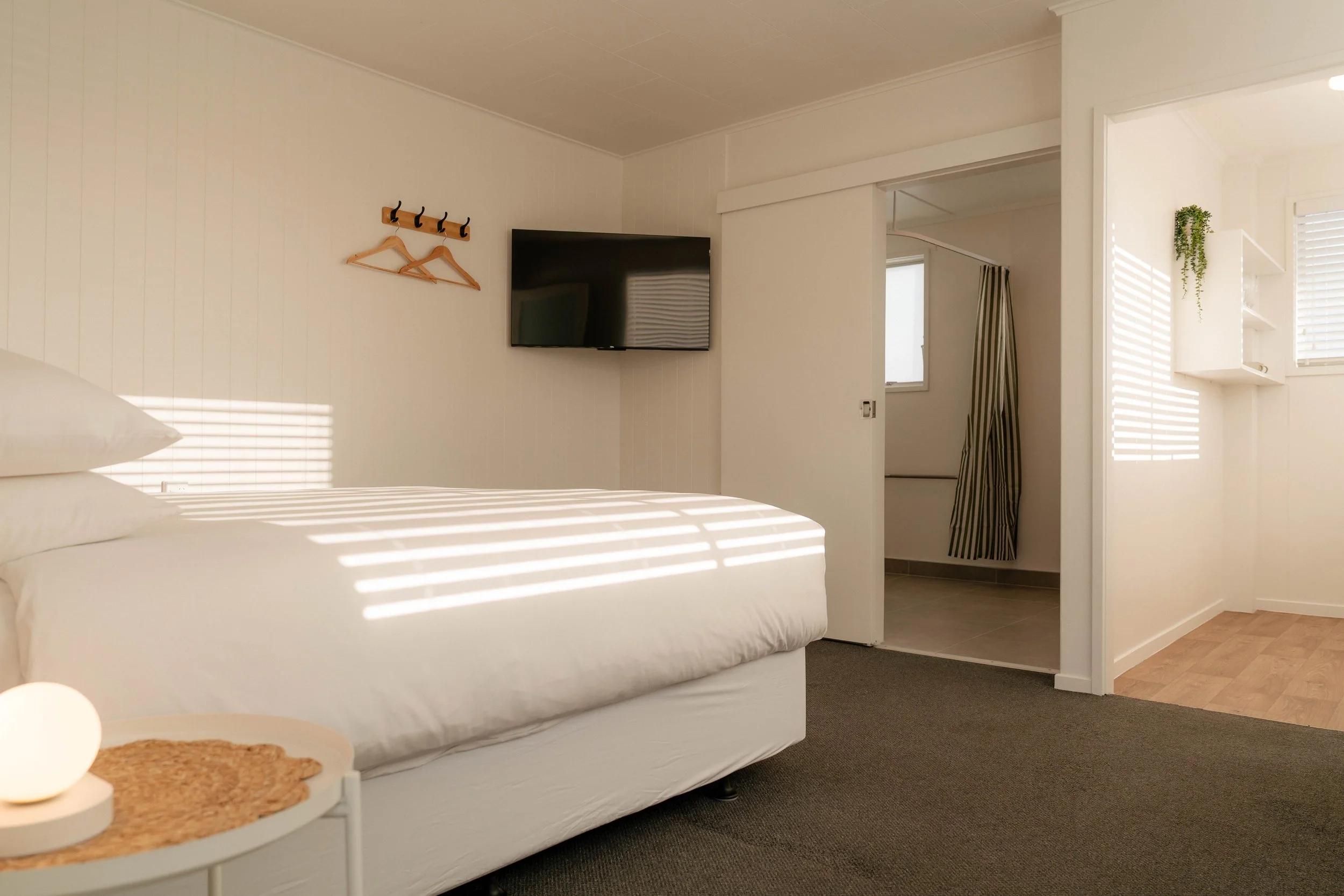 A bedroom with a white bed, a small white side table with a woven placemat, a round lamp, a wall-mounted TV, and a small wooden coat hanger with two hangers on the wall. There is a doorway leading to another room with striped curtains, and sunlight creates shadows through blinds on the walls and bed.
