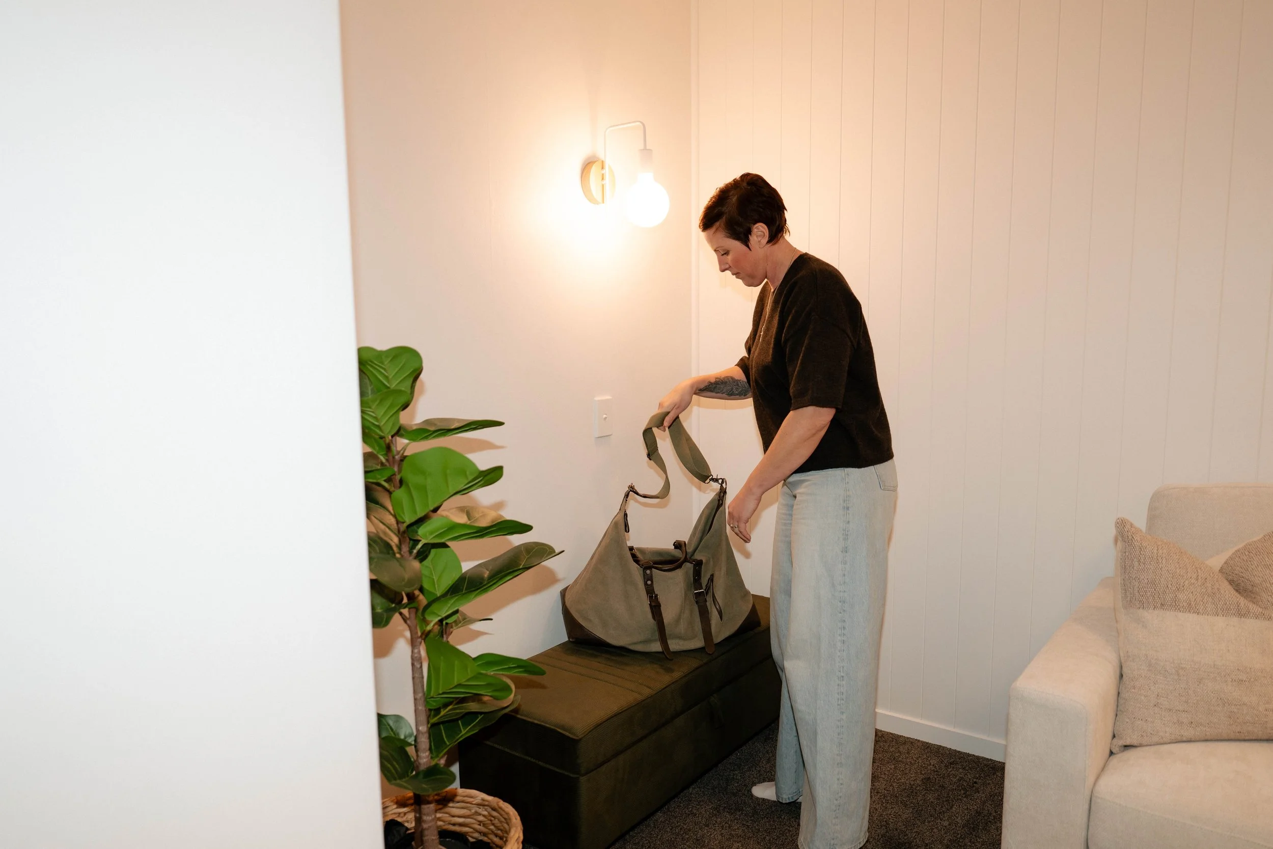 A woman with short dark hair and light skin, wearing a black top and light-colored jeans, is packing belongings into a green tote bag in a cozy, well-lit living room with white vertical panel walls, a beige sofa with cushions, and a potted plant. A wall sconce with a glowing white bulb is mounted on the wall.