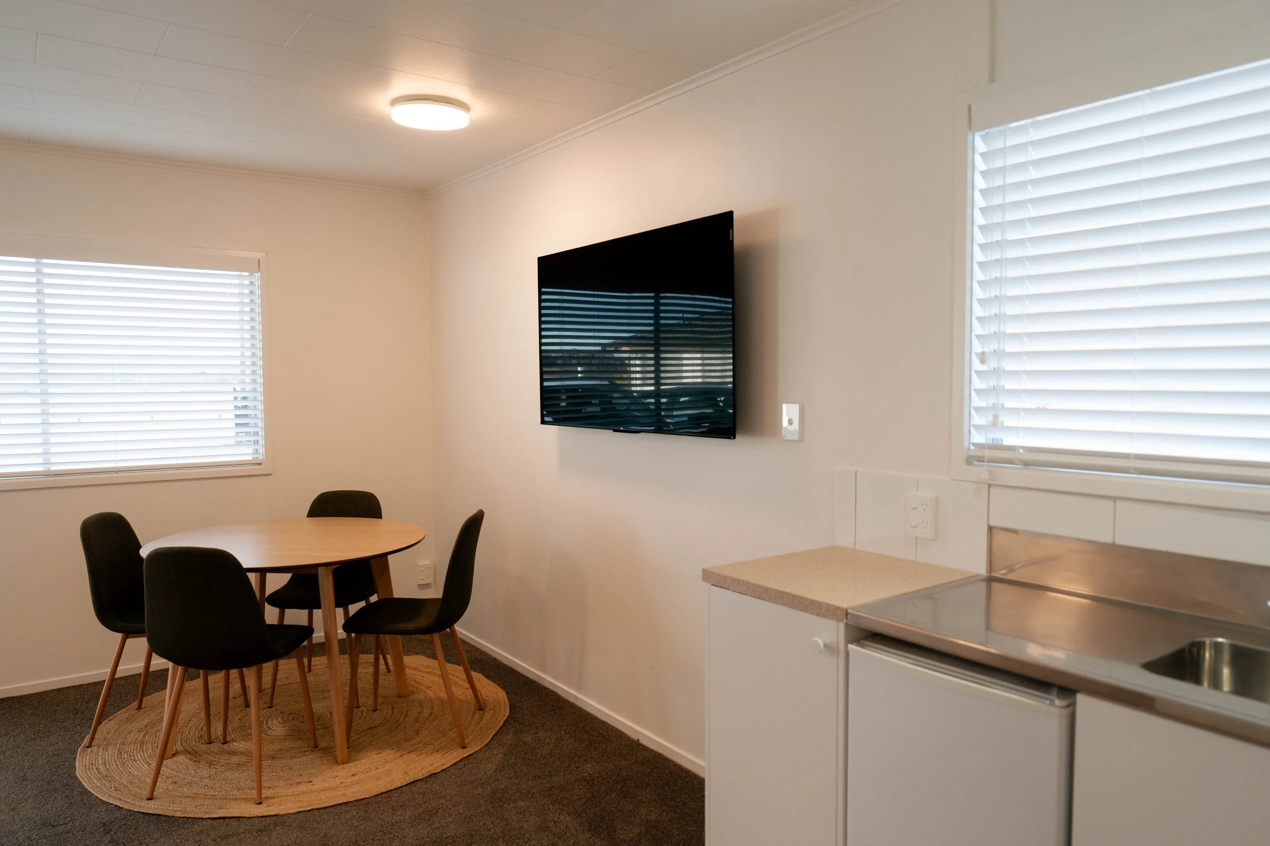A room with white walls, two large windows with blinds, a round wooden table, four black chairs, a wall-mounted flat-screen TV, and a kitchen area with a countertop, sink, and appliances.