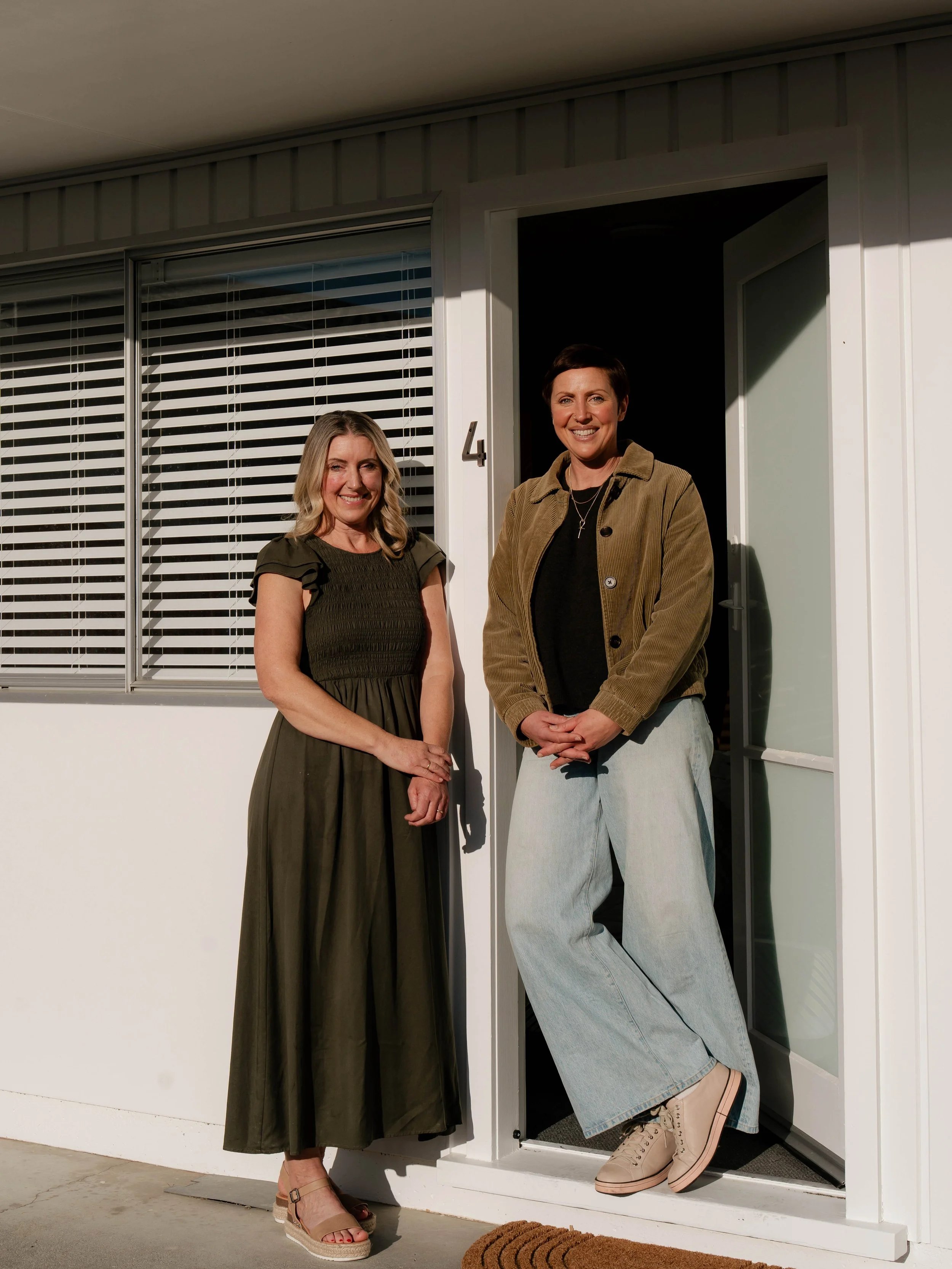 Two women standing outside a house near a door and window with blinds, smiling and posing for a photo.