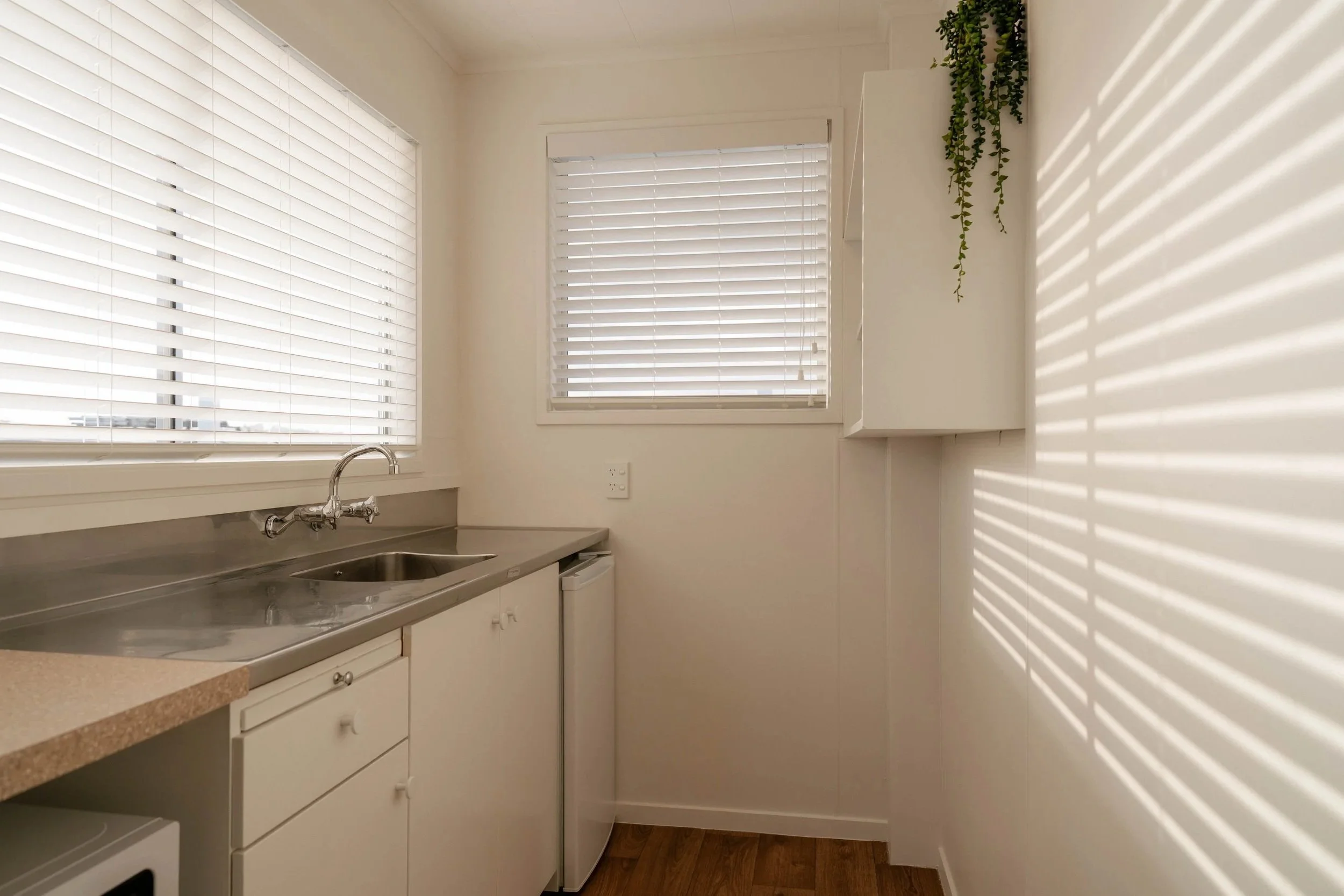 A small kitchen with white cabinets, a stainless steel sink, and hinges. There are two windows with white blinds. Sunlight creates shadow patterns on the wall. A small green plant is on top of the cabinet.
