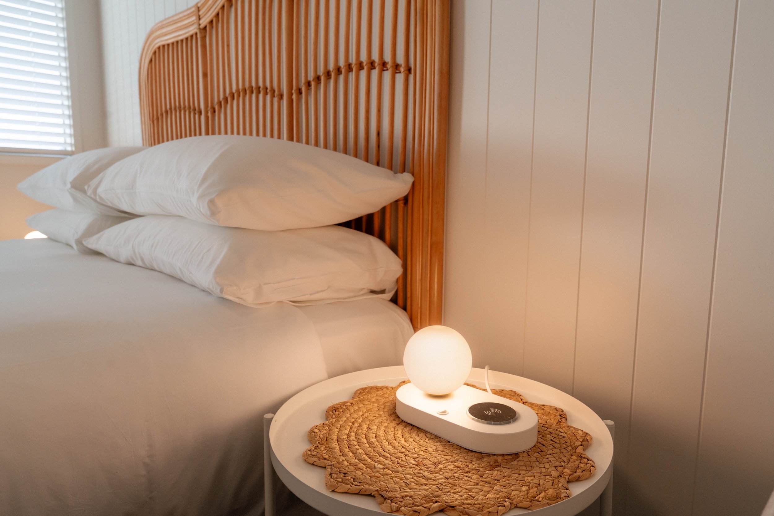 A cozy bedroom with a wooden headboard, white bedding, and a white nightstand. The nightstand holds a round woven mat with a modern white lamp and a black wireless charging pad. Natural light filters through a window with white blinds.