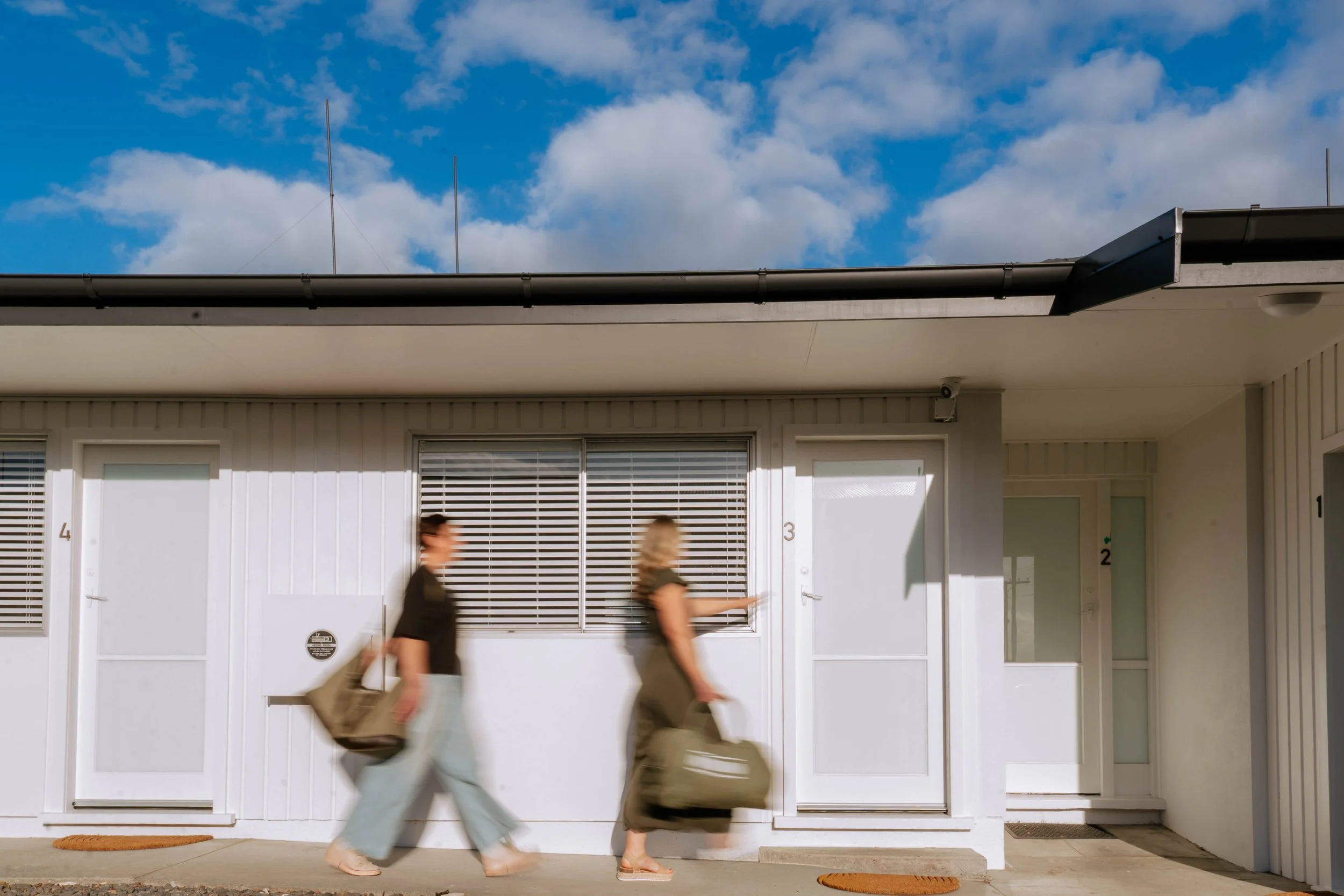 Two people walking past a modern apartment building with gray siding and white doors, numbered 4 and 3, under a cloud-filled blue sky.
