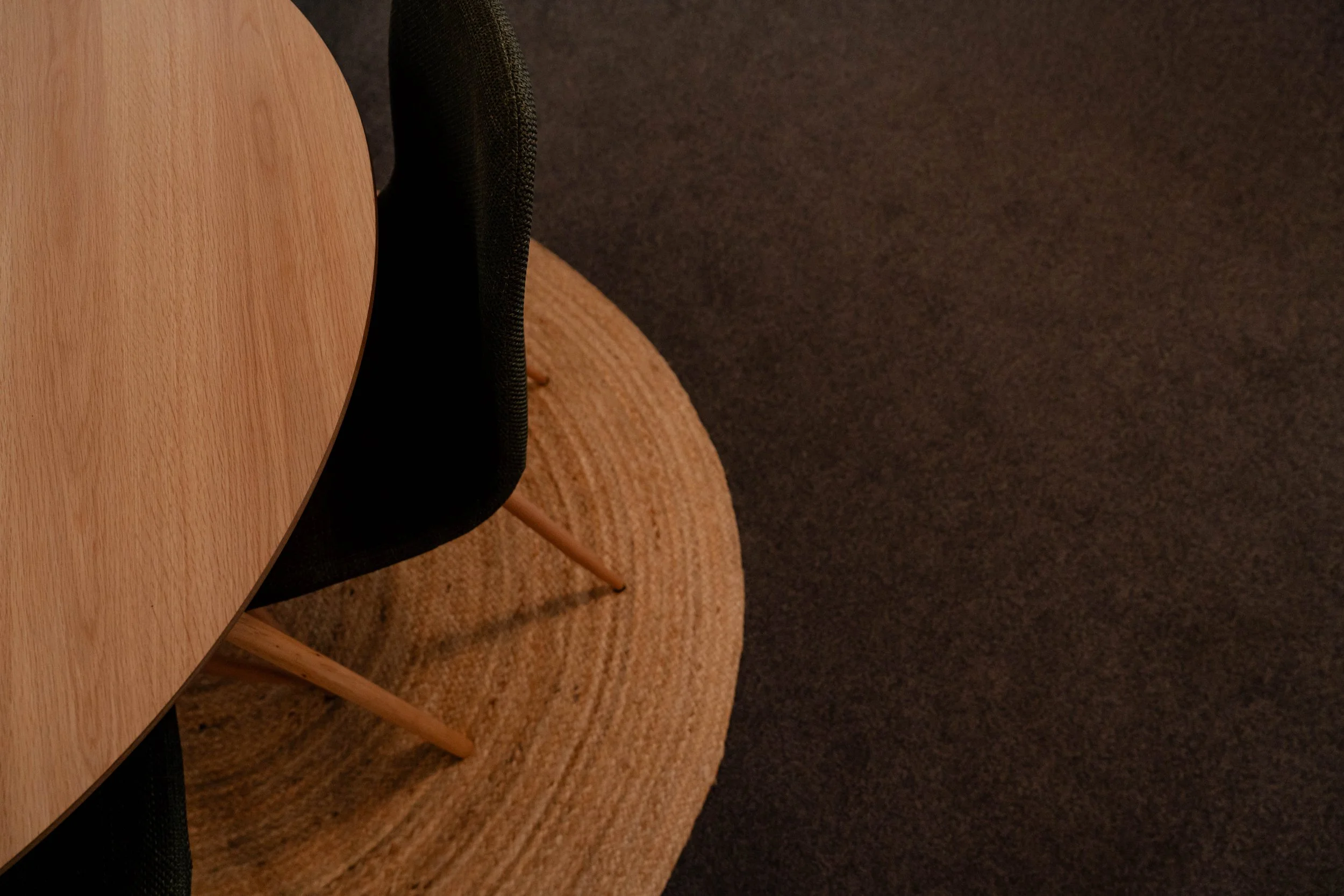 Part of a round wooden table, a black upholstered chair, and a wooden chair on a textured beige rug, with a dark carpeted floor in the background.