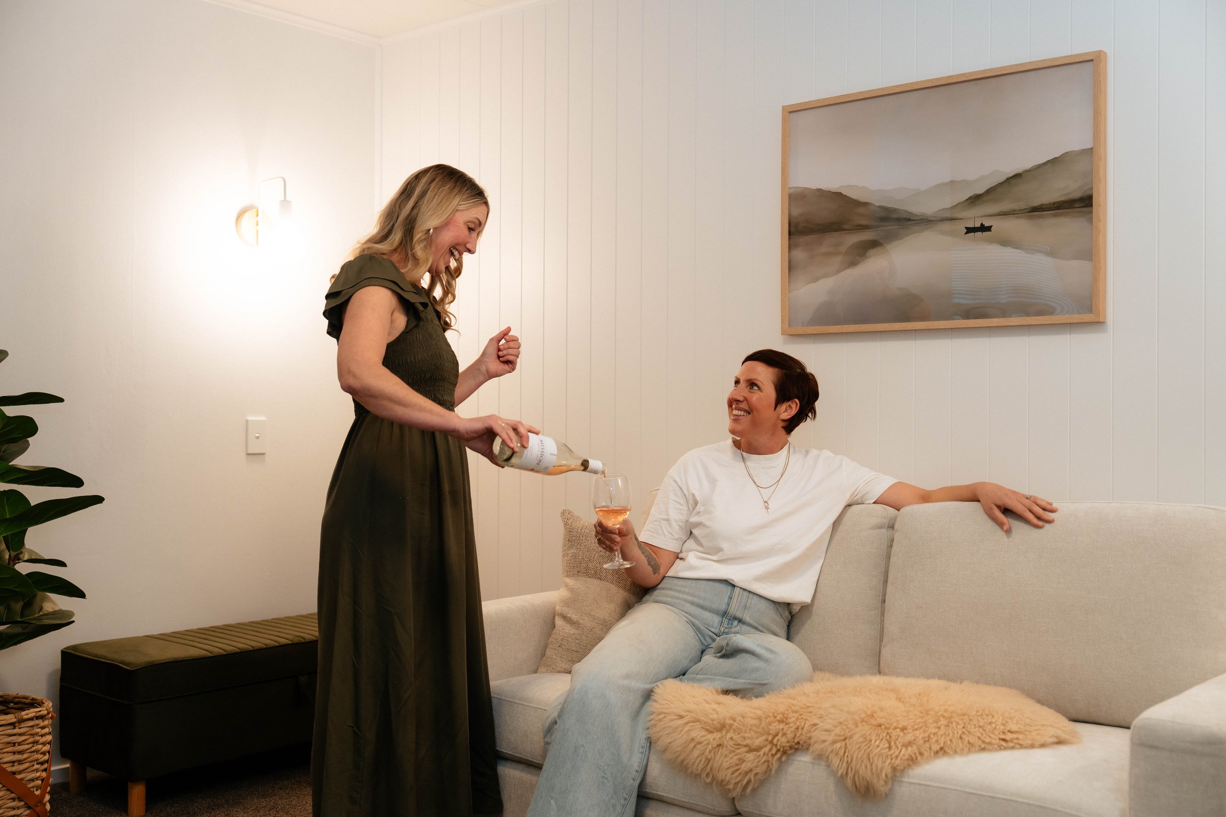 A woman standing pouring wine into a glass held by a woman sitting on a sofa, in a cozy living room with a framed landscape picture on the wall and warm lighting.