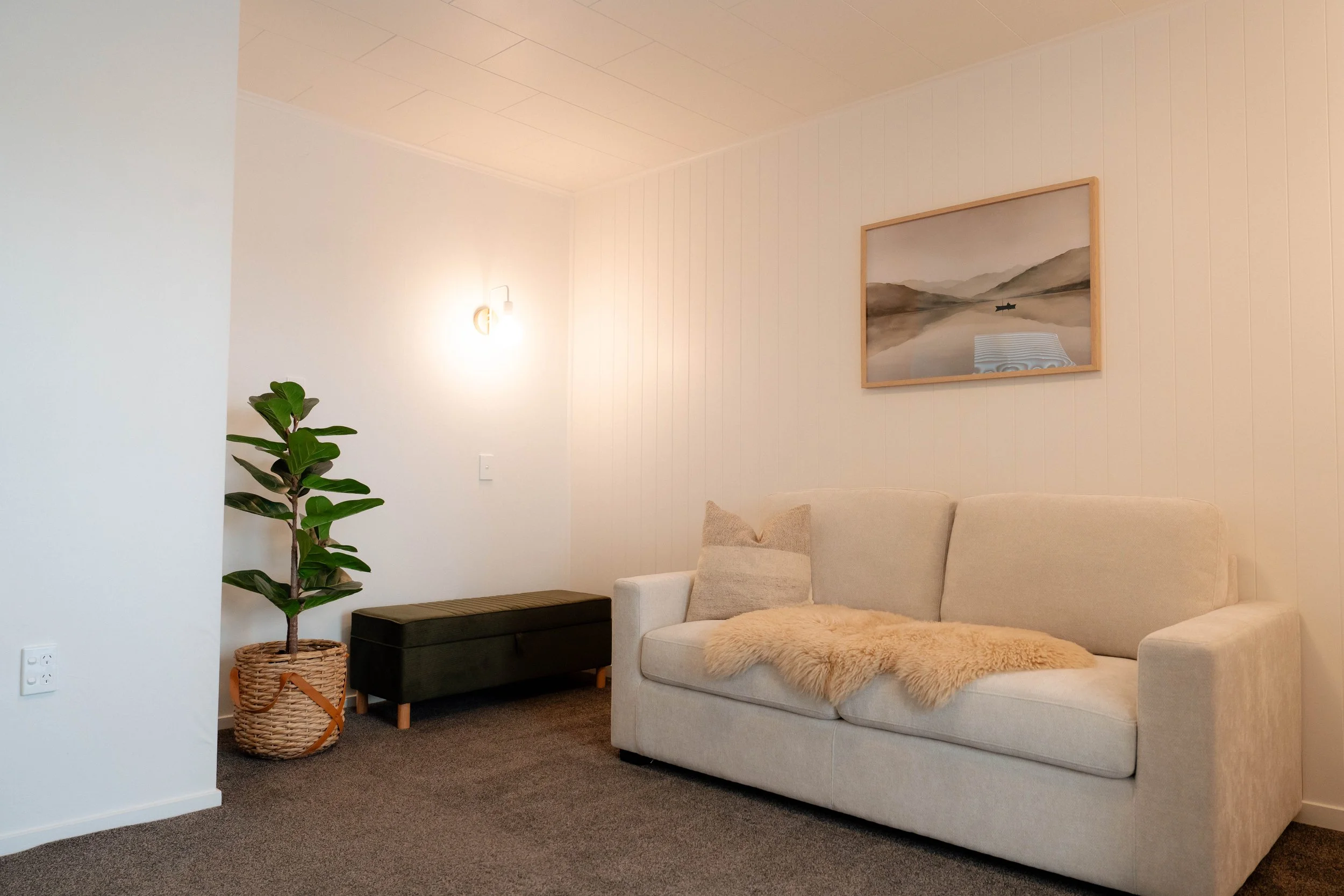 Living room with a beige sofa, pillows, a blanket, a black storage bench, a potted plant, a wall-mounted light, and a landscape painting on a cream-colored wall.