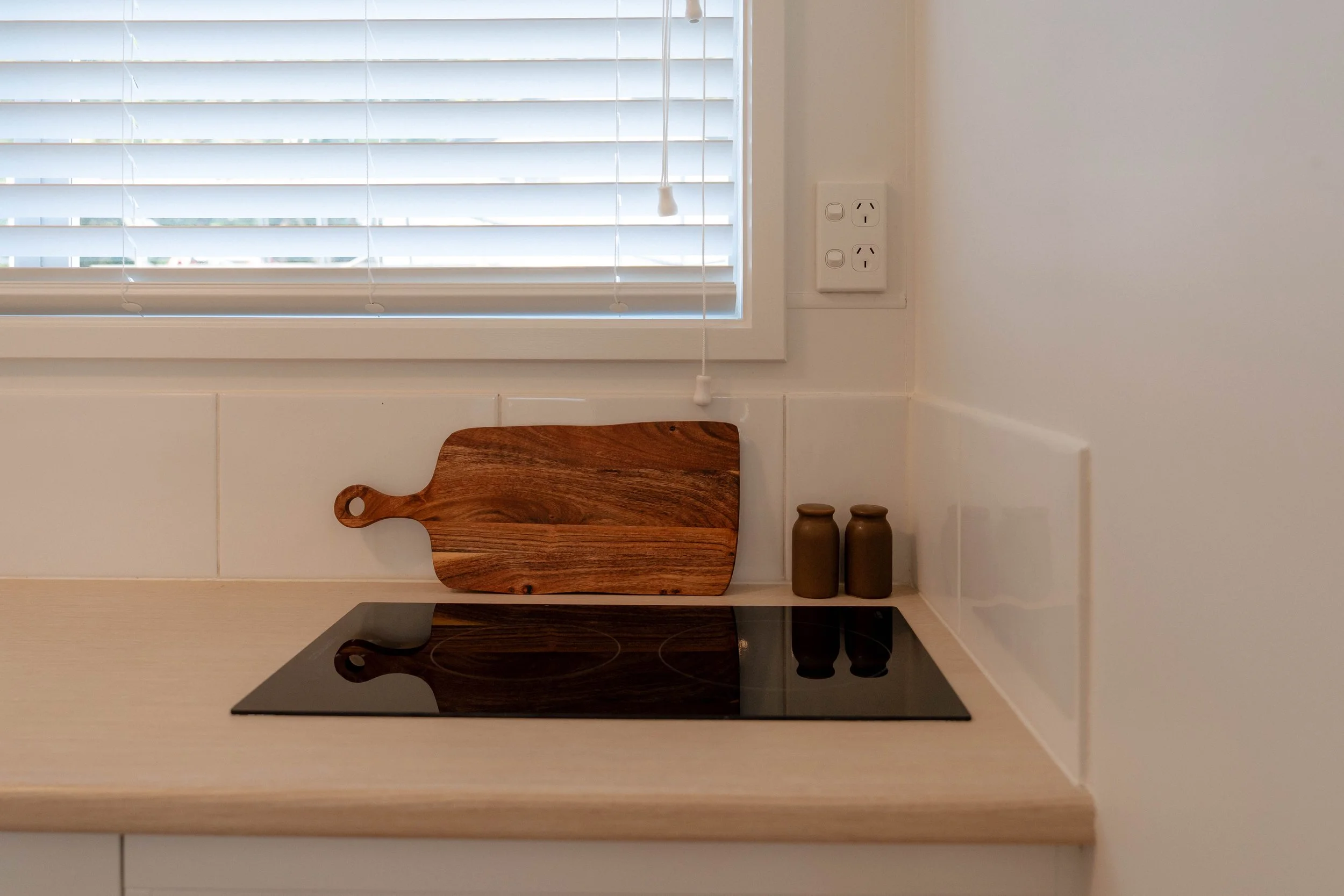 A kitchen window with white blinds, a wooden cutting board, two small decorative jars, and a black stovetop underneath.