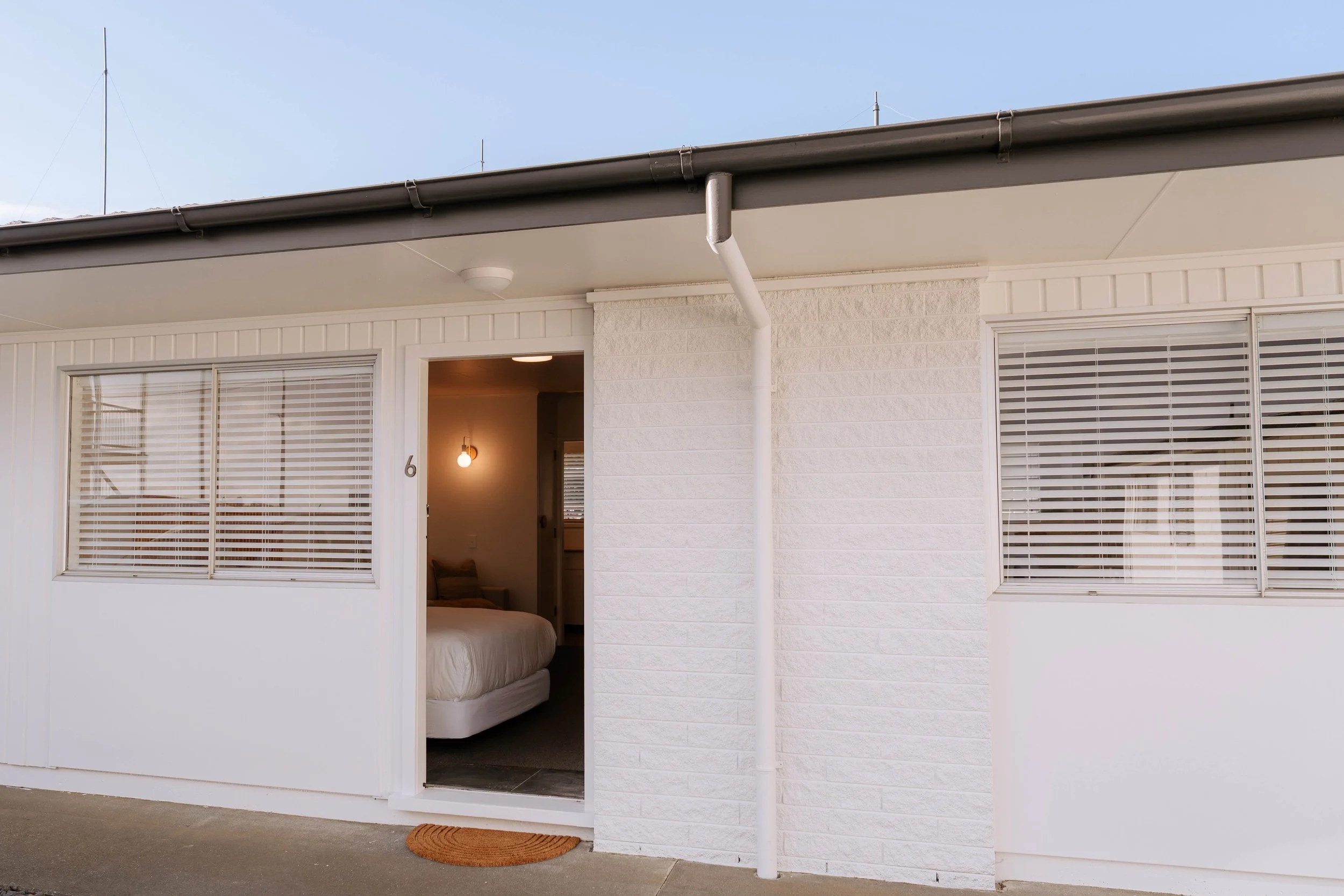 The exterior of a house with a sliding door open revealing a bedroom inside. The house has white walls, two large windows with horizontal blinds, and a brown doormat outside the entrance. The sky is clear and partly cloudy.