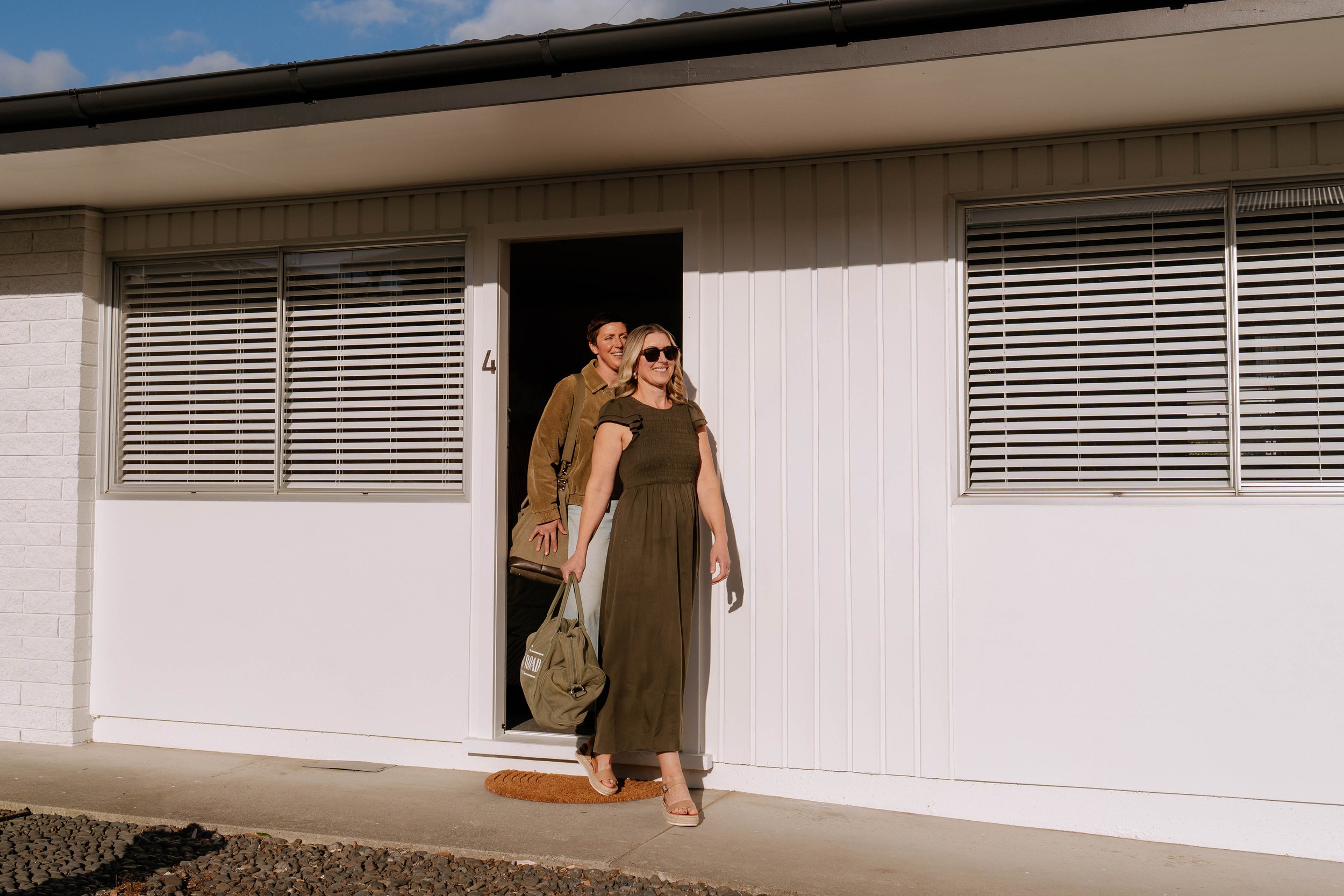 Two women are standing in the doorway of a white building with horizontal blinds on the windows. One woman is wearing dark sunglasses and a long dress, while the other woman is smiling behind her, wearing a casual shirt. They appear to be happy.