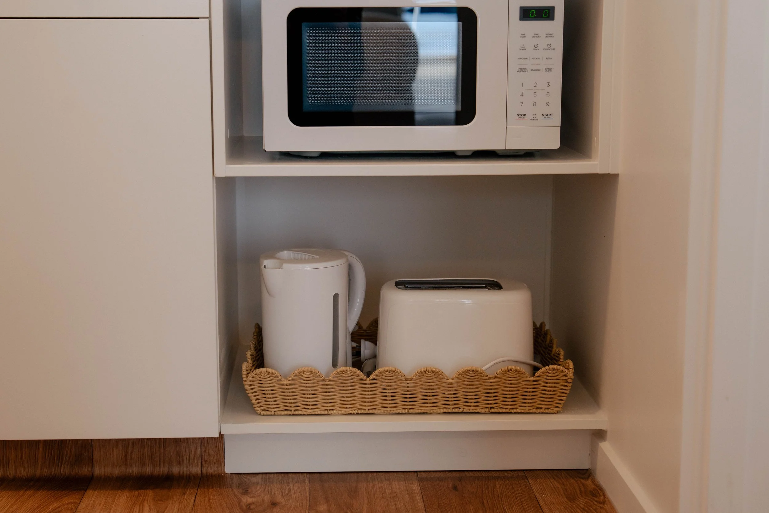 An open kitchen cabinet shelf showing a microwave oven on top, a white electric kettle and a toaster in the middle, and a woven basket at the bottom. The cabinet has a white interior and wood flooring is visible below.