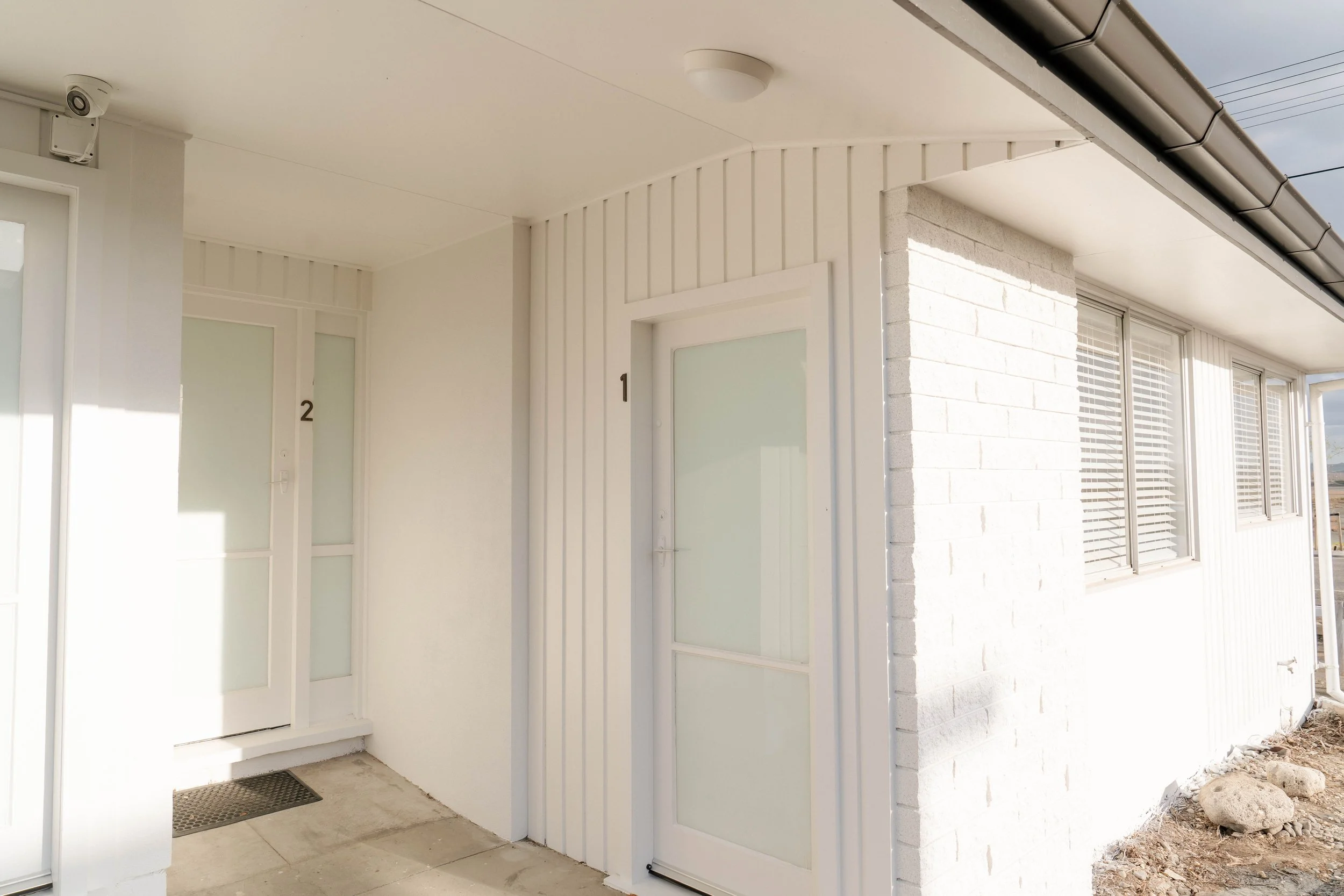 Front porch of a modern white brick house with glass patio doors, a white door numbered 1, and a side wall with vertical siding. There's an overhang with a light fixture and a security camera, and a gutter along the edge of the roof.