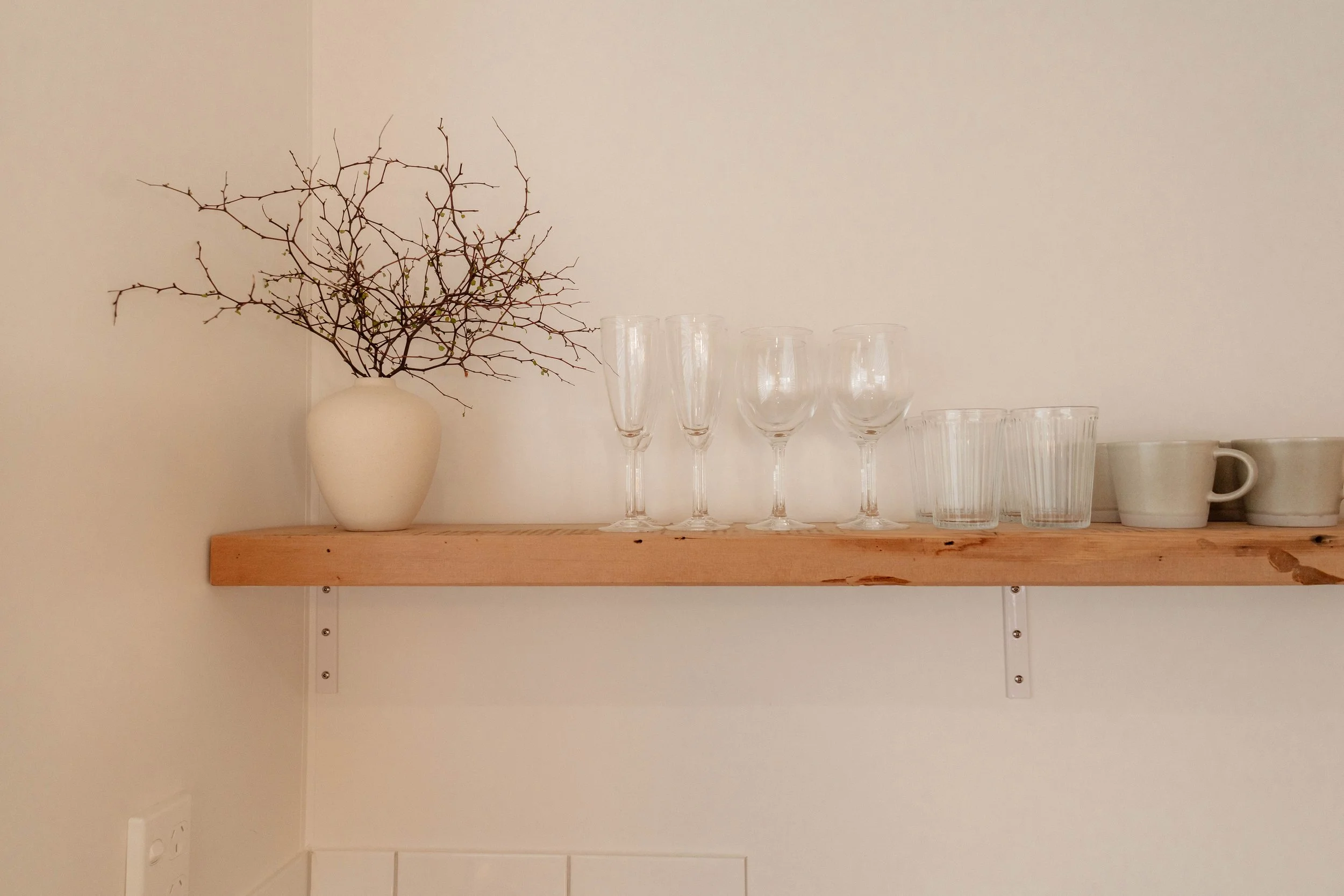 A wooden shelf with a white vase holding a bare, leafless branch, five empty champagne flutes, three empty glasses, and two coffee mugs against a plain light-colored wall.