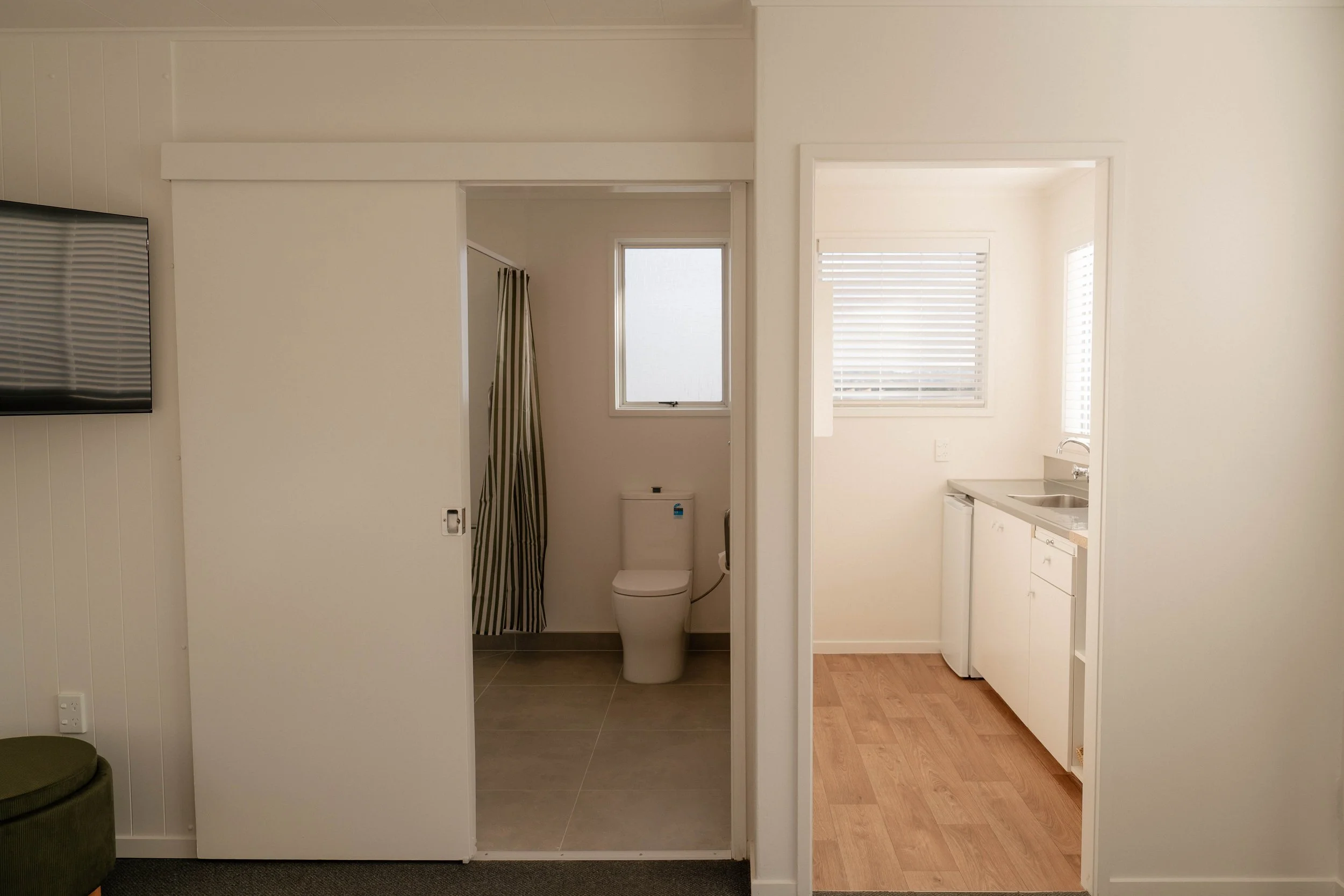 Image shows a split view of a small bathroom with a toilet and a shower on the left, and a kitchen with a sink and white cabinets on the right. There are windows with blinds on both sides, and the flooring varies between tile and wood.