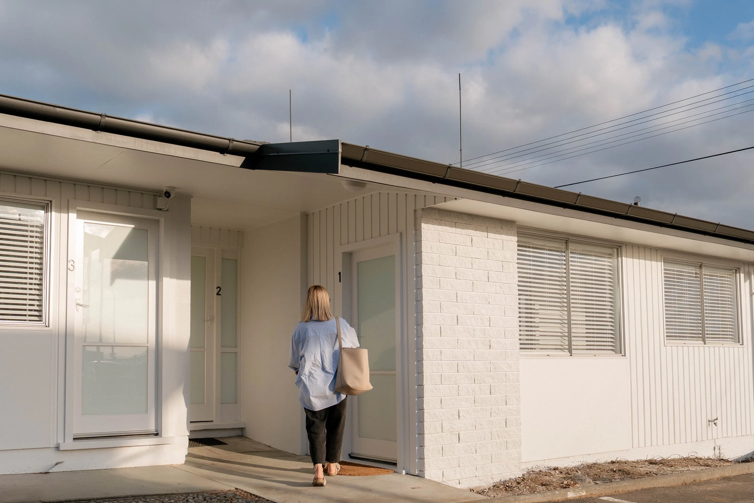 A woman with blonde hair wearing a white shirt and black pants carrying a beige tote bag walking towards a white modern house with large windows.