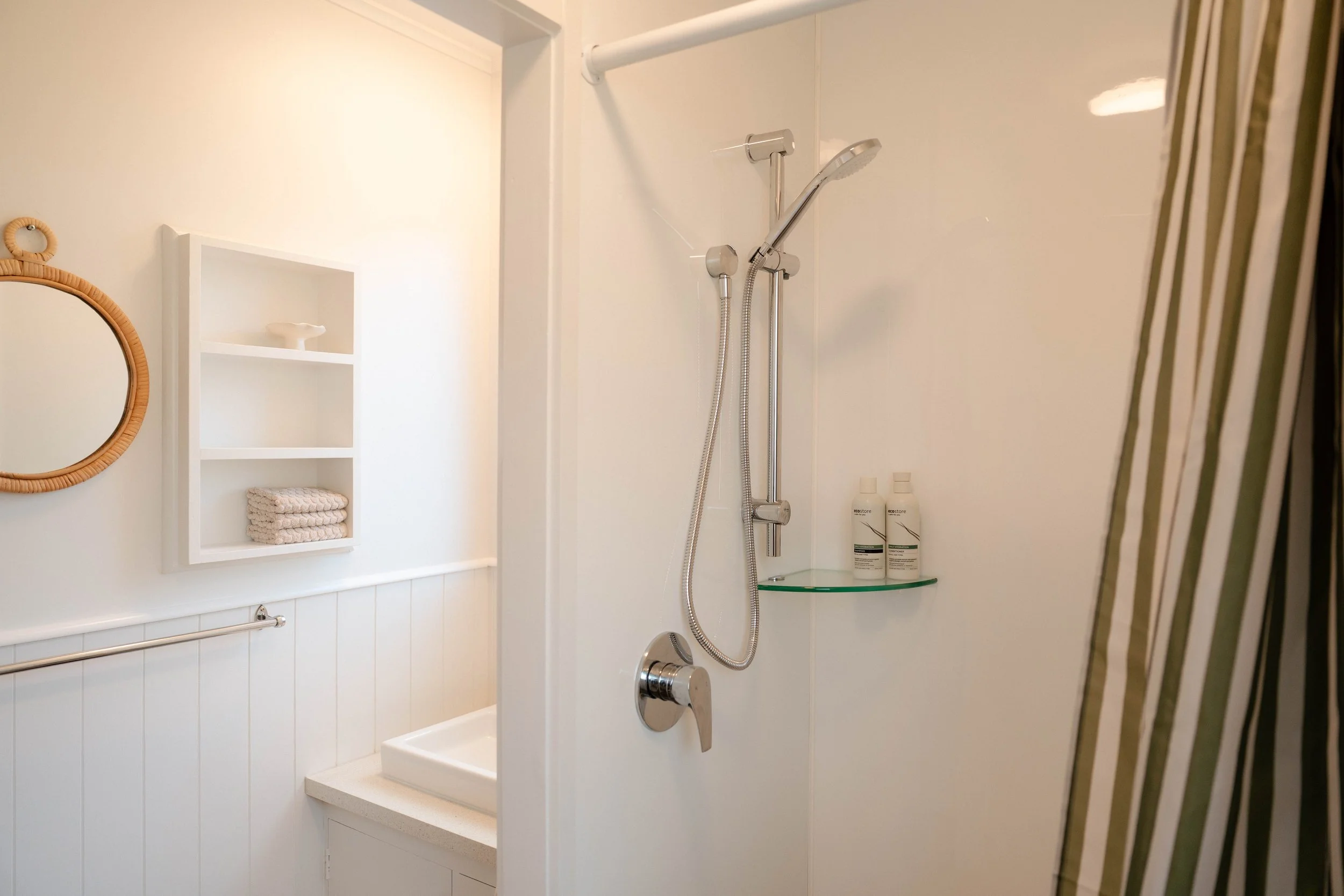 A small shower area with a handheld shower head, soap, and shampoo bottles, a shelf with folded towels, and a round mirror with a wicker frame on the wall.