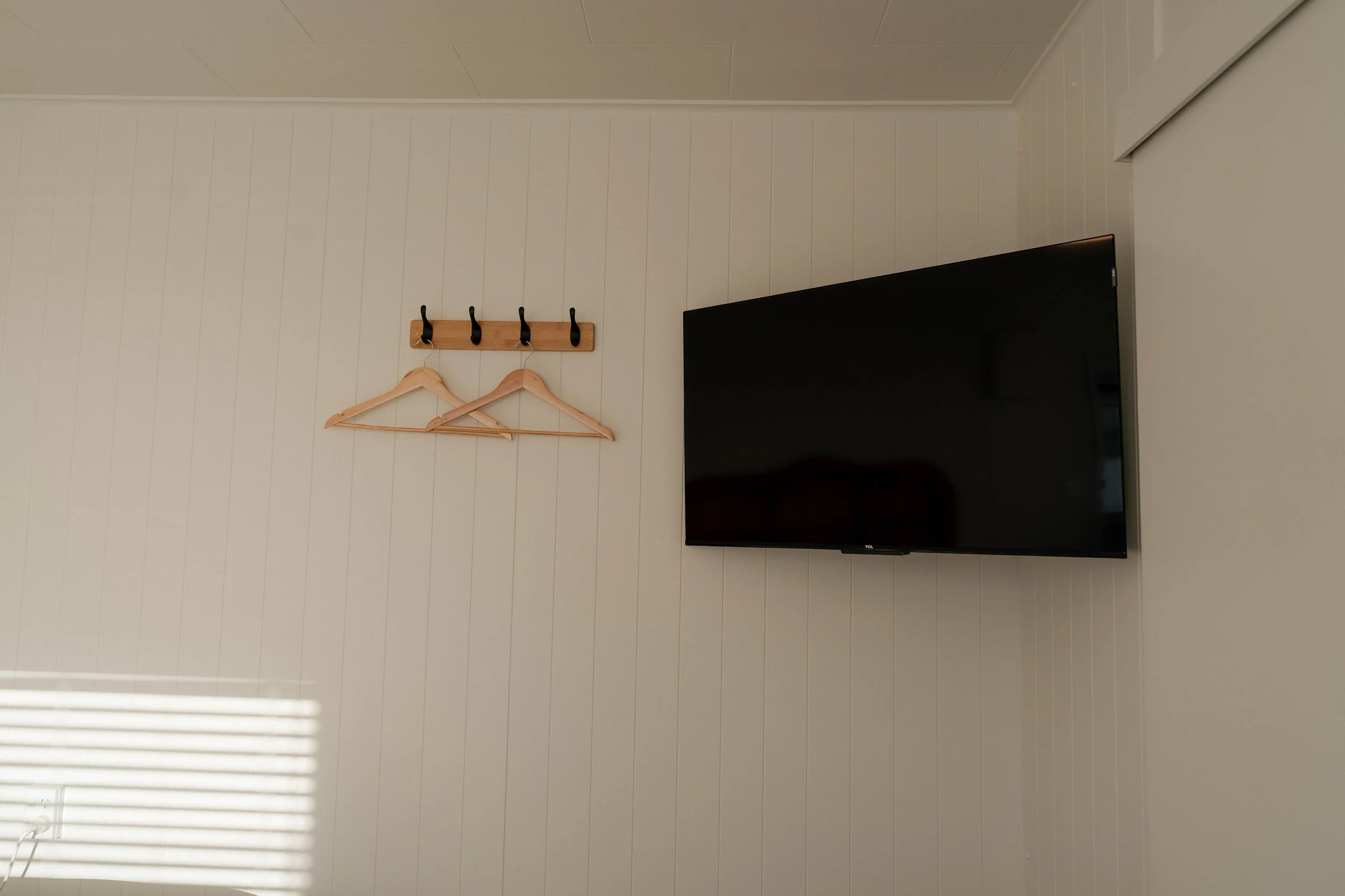 A wall with vertical white paneling, a wooden coat rack with four black hooks, two wooden hangers on the hooks, a large flat-screen TV mounted on the right side, and a partially visible window with blinds casting shadows at the bottom left.