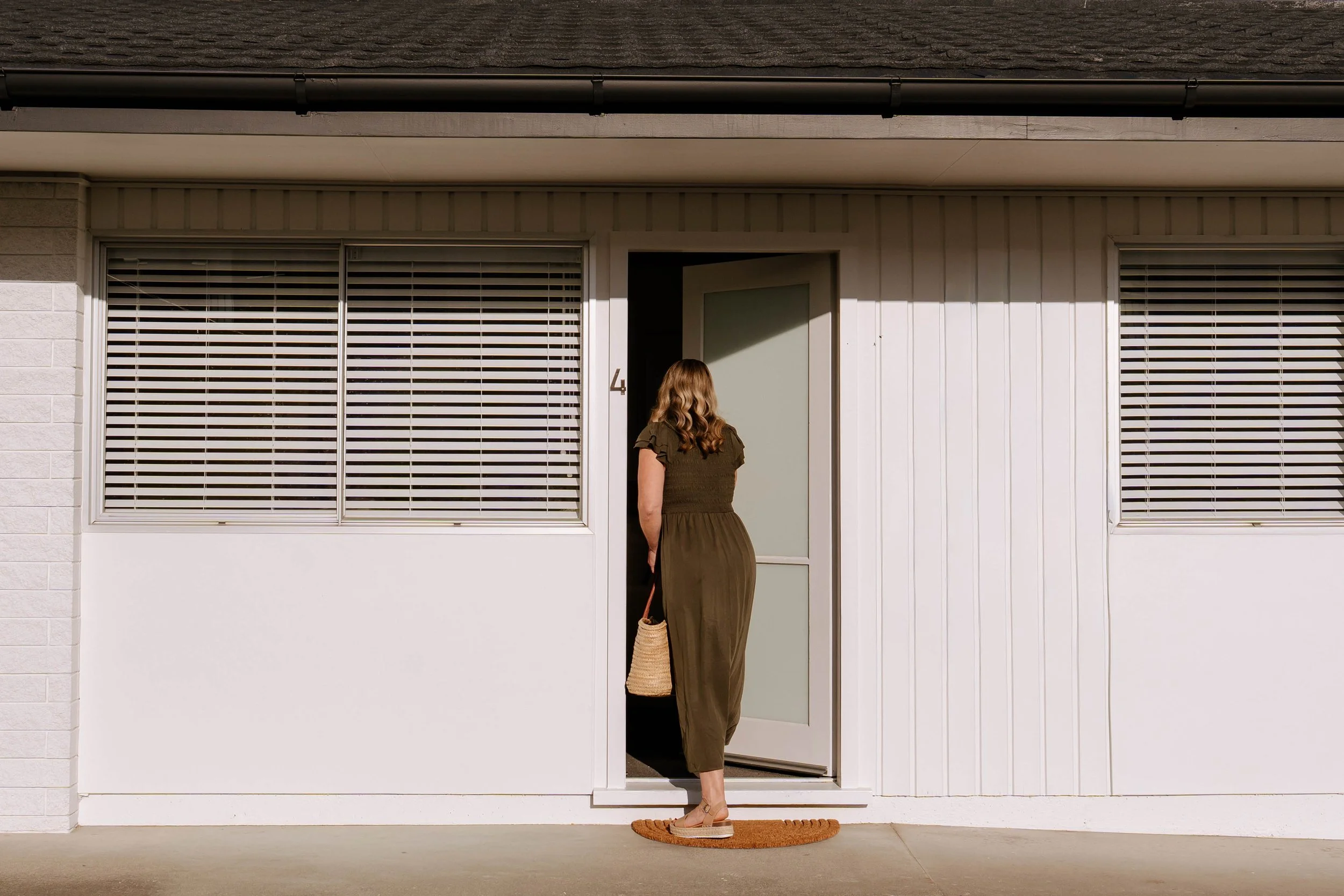 A woman with wavy brown hair wearing an olive green dress and sandals stands outside a modern house, holding a straw bag, with her back to the camera, near a slightly open door and two large windows with horizontal blinds.