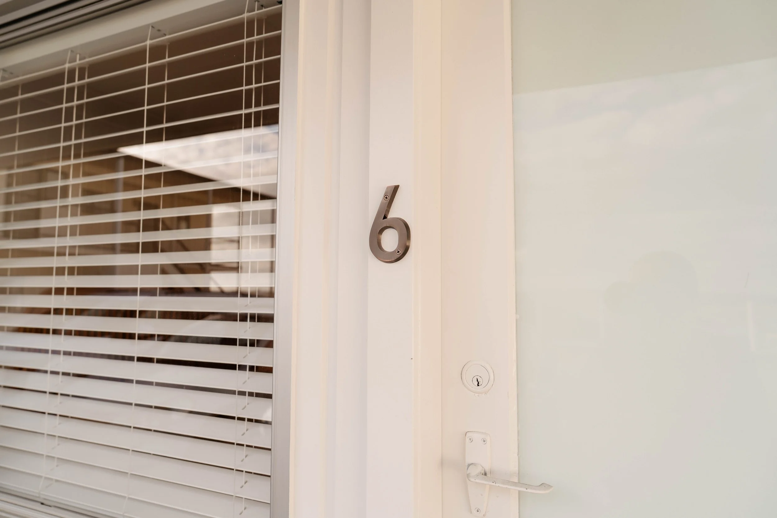 Close-up of a white window with horizontal blinds and a inch number '6' metallic plaque in a building's interior.