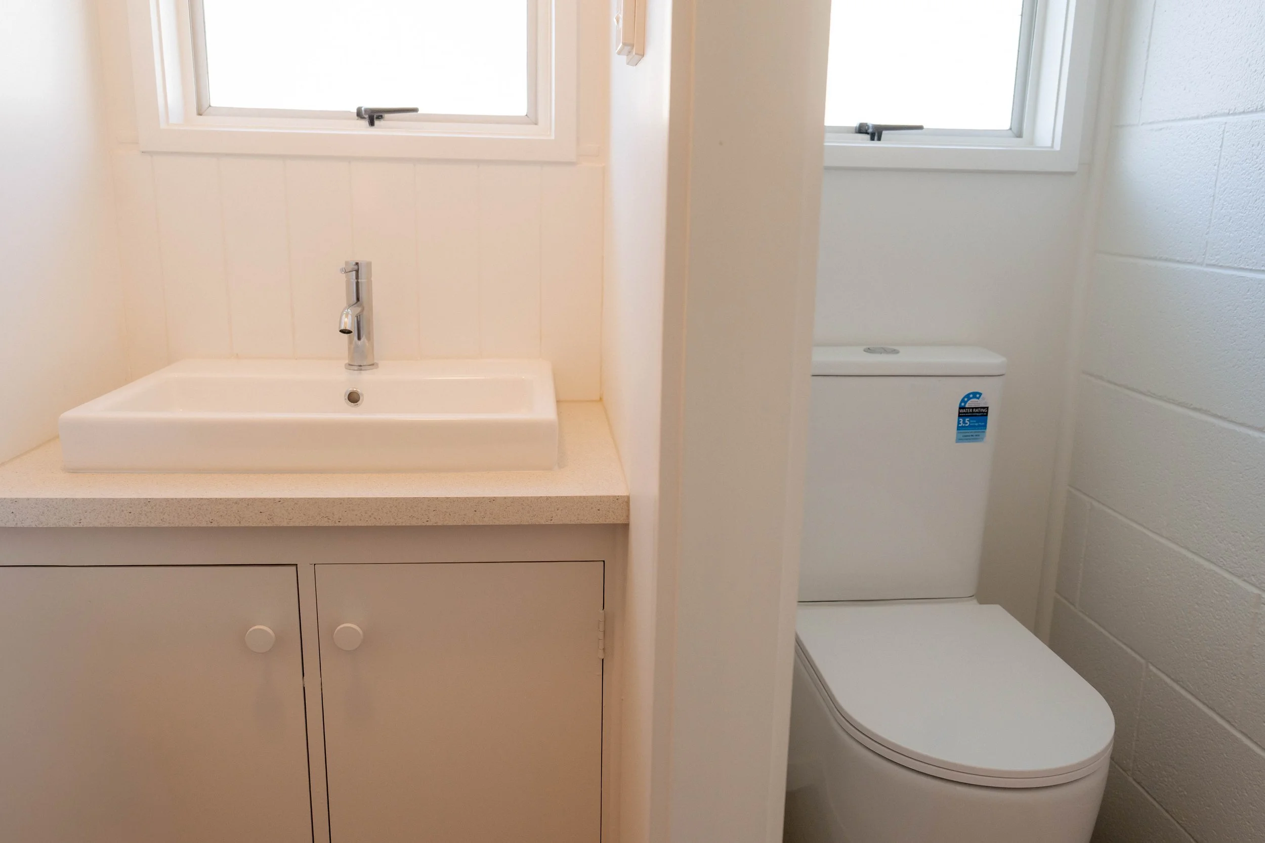 Bathroom with white sink and faucet, small cabinet below, toilet with a water rating sticker, two windows with white frames, light-colored walls and countertops.