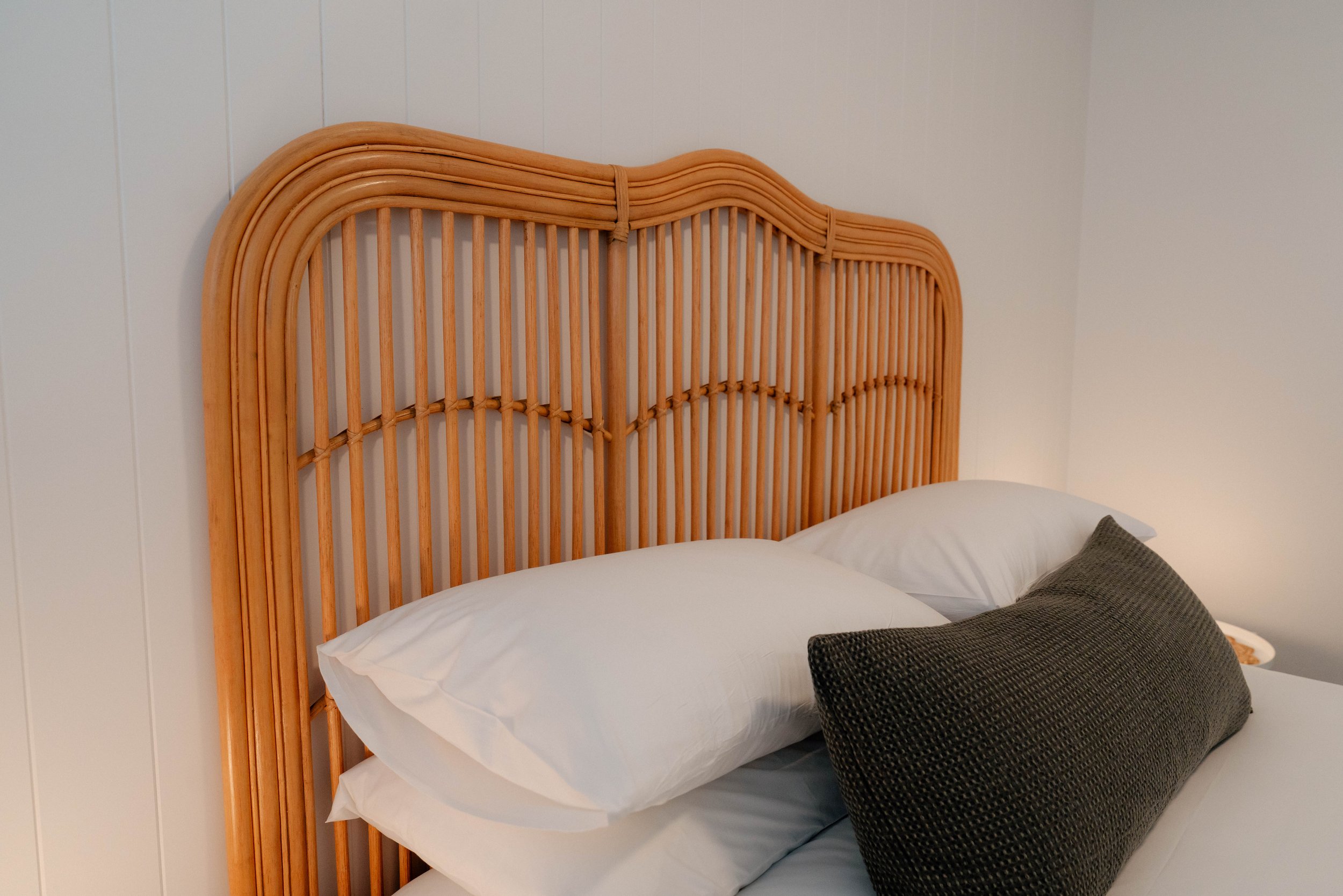 A bed with a rattan headboard, white pillows, and dark textured pillows against white paneled walls.