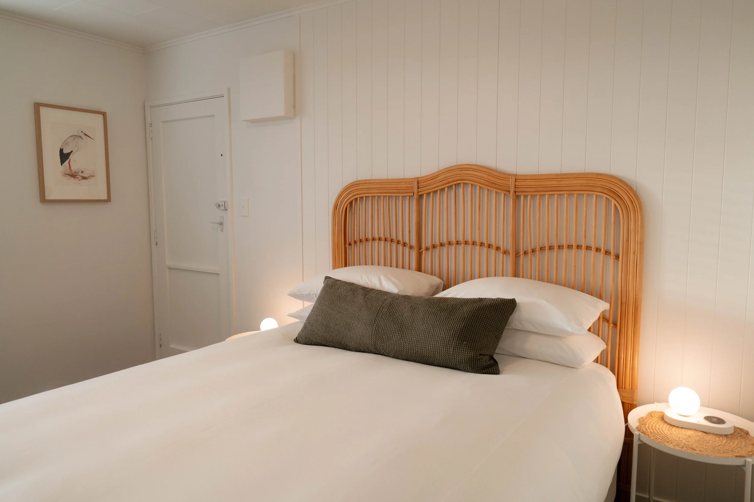 A bedroom with white walls, a rattan headboard, white bedding, multiple pillows, and a framed bird illustration on the wall. A nightstand with a small lamp and decorative items is beside the bed.