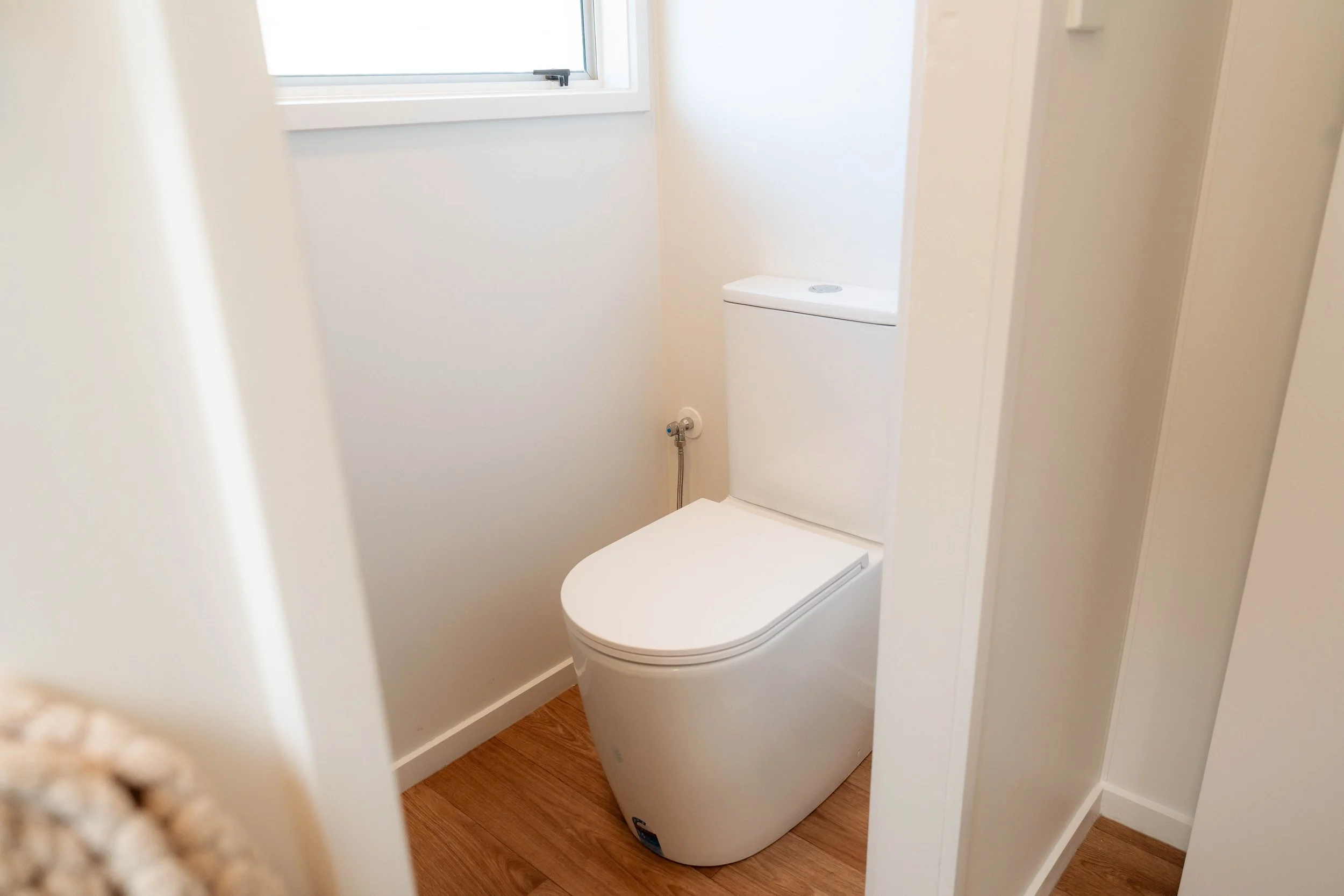 A modern white toilet with a rectangular tank, placed in a small, white bathroom with a window and light wood flooring.