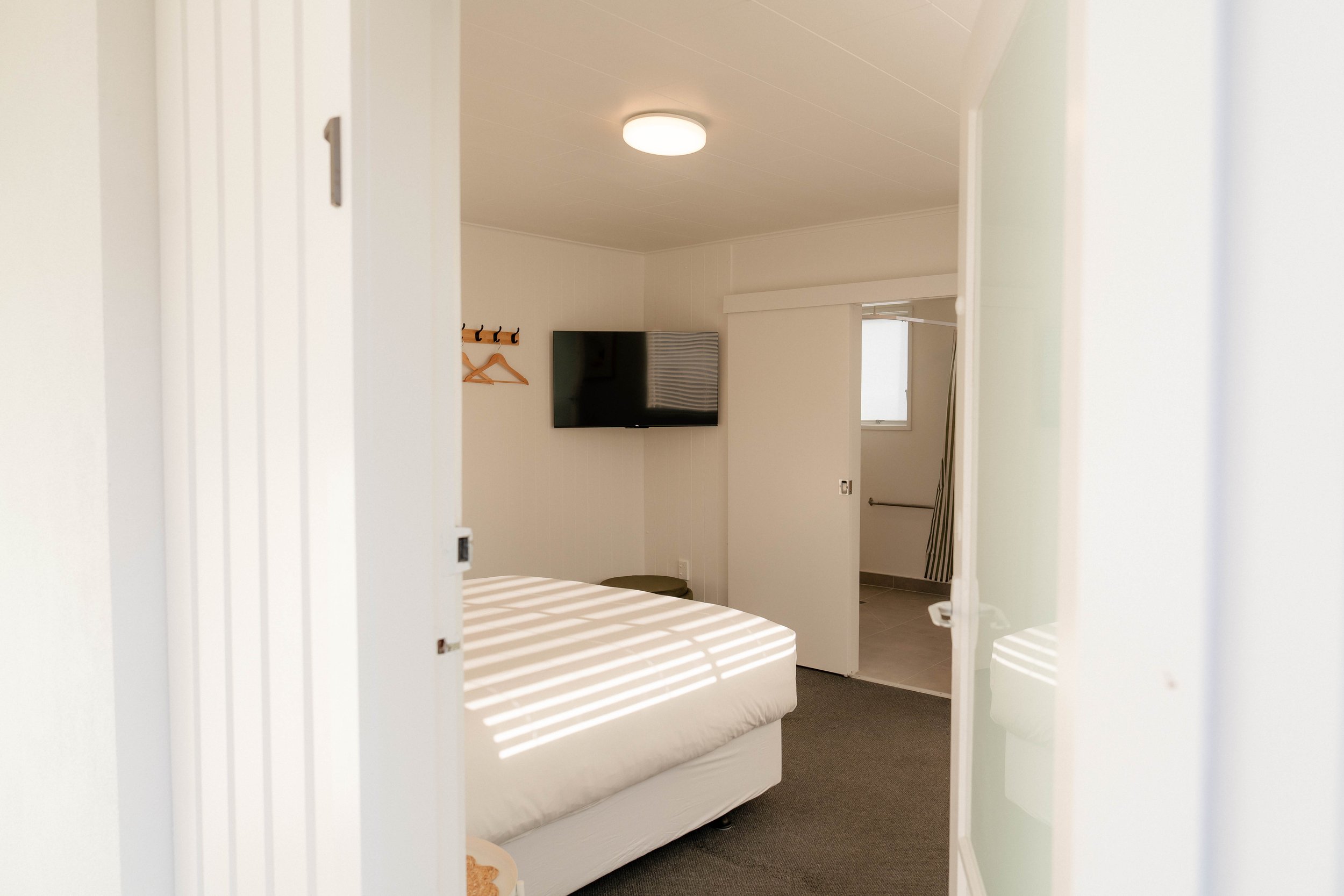 A room viewed through a slightly open door, showing a bed with a striped bedspread, a wall-mounted flat-screen TV, a small window, and a coat rack with two hangers.