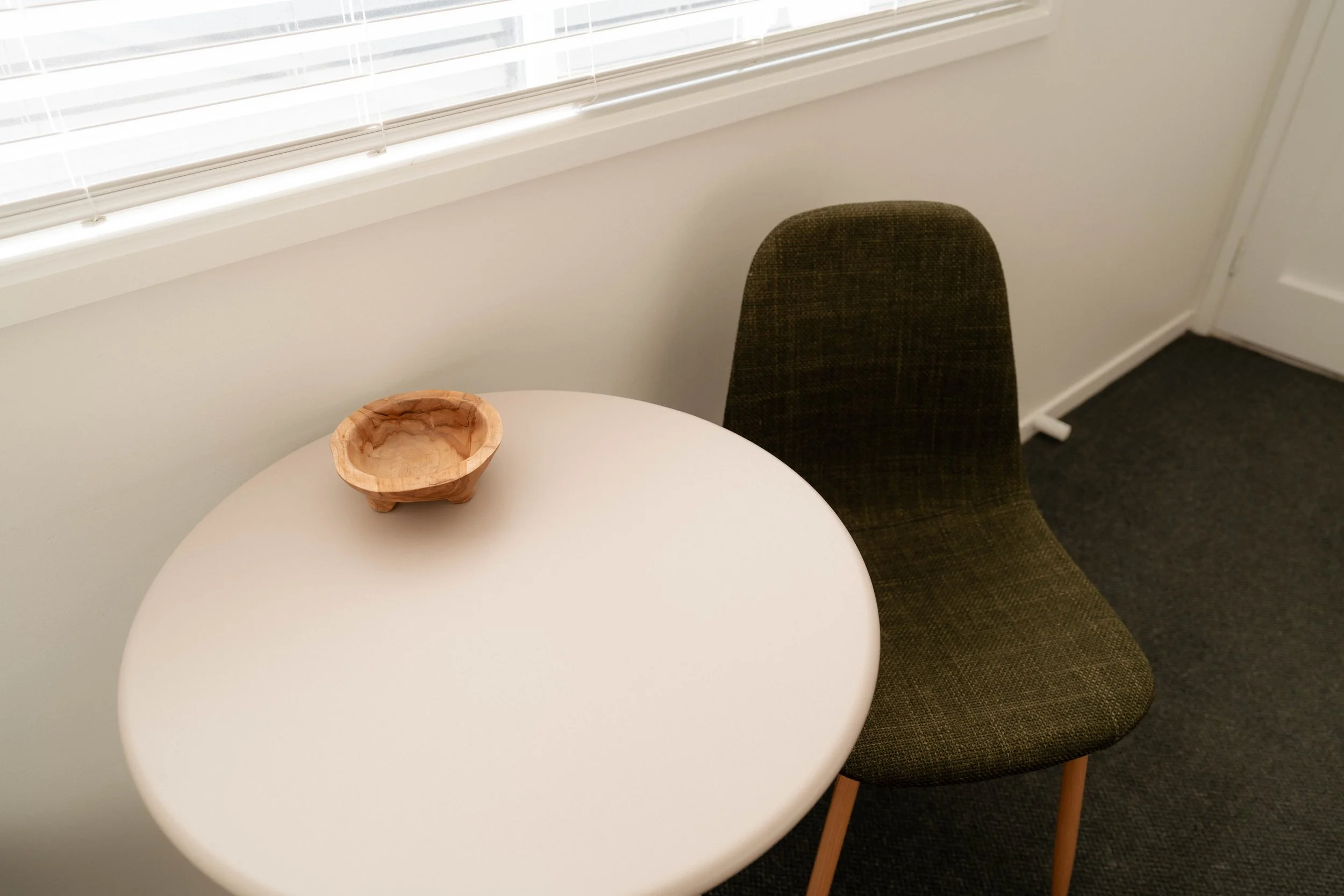 A white round table with a small wooden bowl on top, a brown upholstered chair beside it, a window with blinds in the background, and a white wall and door.