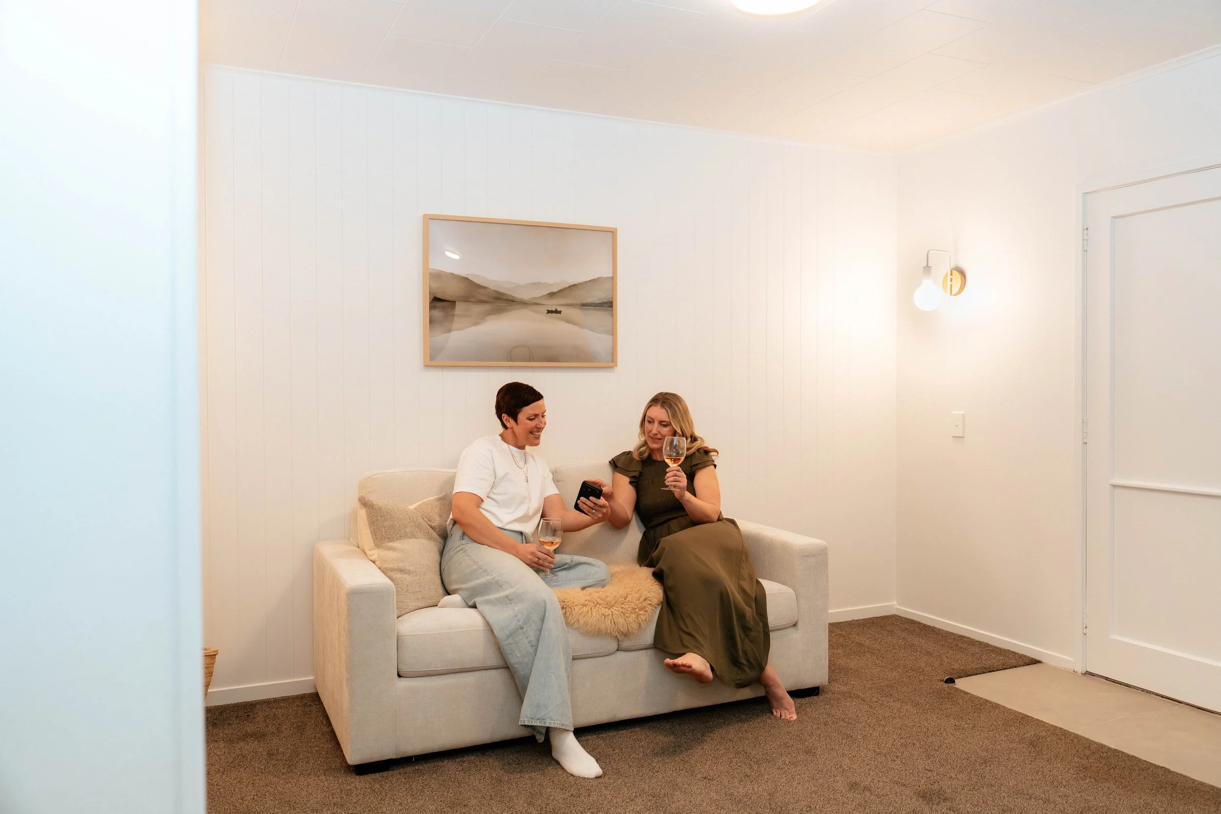 Two women sitting on a light-colored sofa in a cozy, brightly lit room, engaged with a smartphone and holding glasses of rosé wine. One woman has short dark hair and is wearing a white shirt and light jeans, while the other has long blonde hair and is wearing a dark green dress. The wall behind them is decorated with simple artwork featuring mountains and water, and there is a modern wall sconce light and a door to the right.