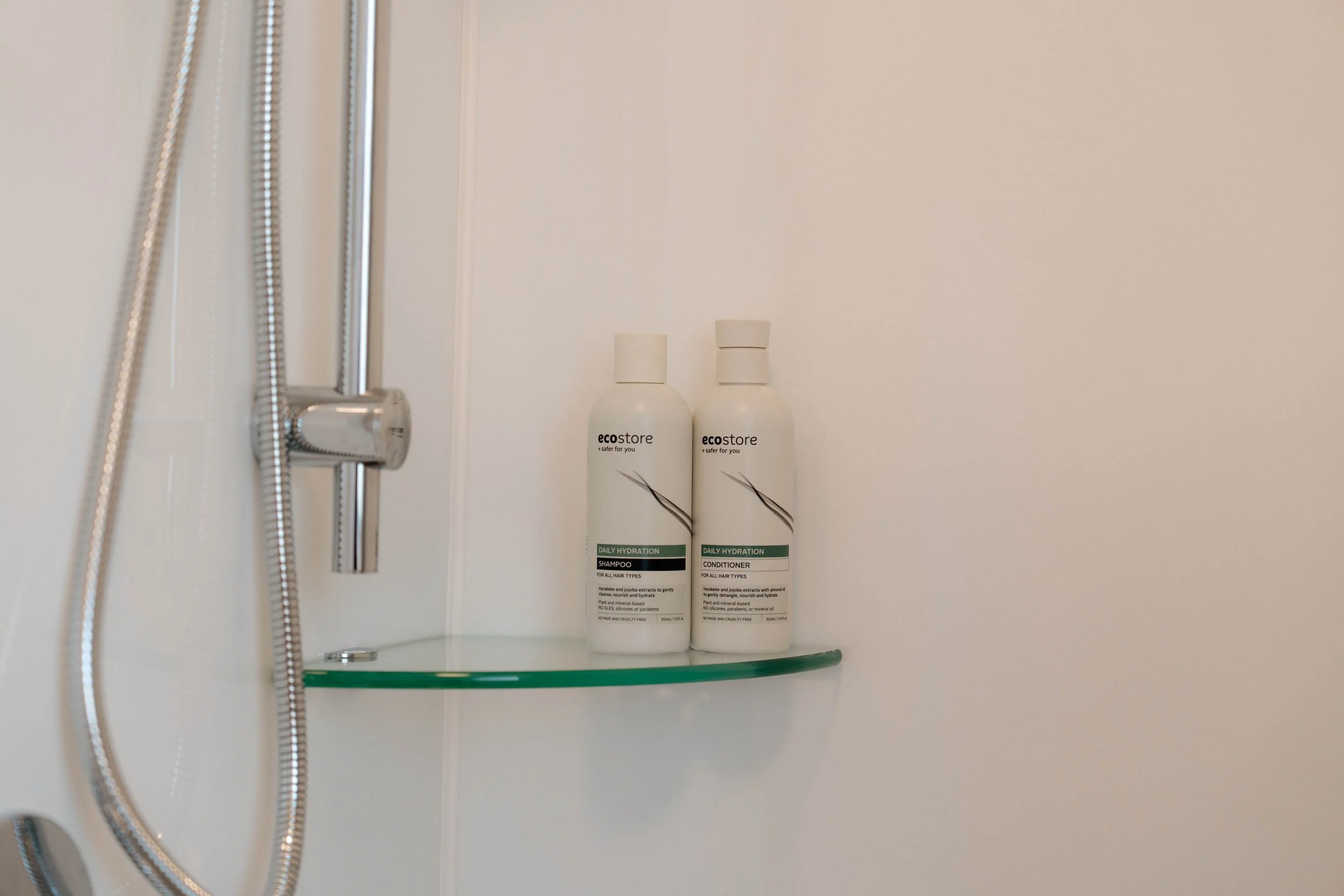 Two white bottles of eco store daily hydration shampoo and conditioner with black and grey text, placed on a glass shelf inside a shower, with a metal shower head and hose visible on the left.