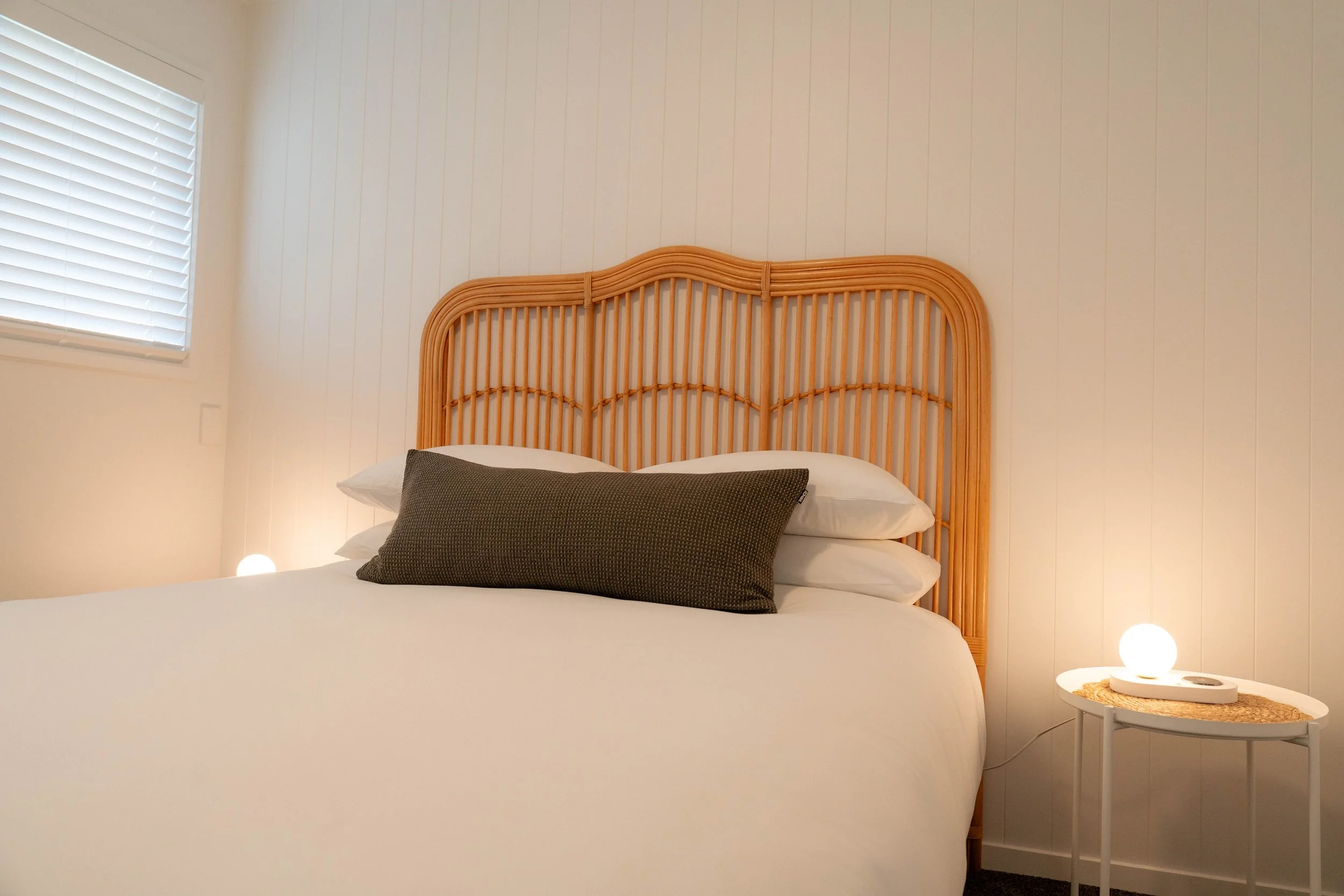 A bedroom with a wooden headboard, white walls, a window with closed blinds, white bedding, a dark pillow, and a small side table with a round top and a lamp.