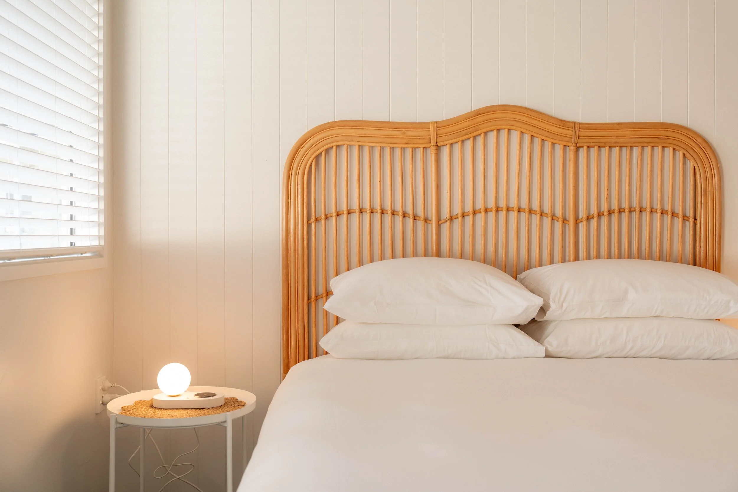 A bedroom corner with a bed featuring a rattan headboard, white bedding, a small white side table with a woven placemat, a white spherical lamp, and white window blinds. The room has cream-colored walls.