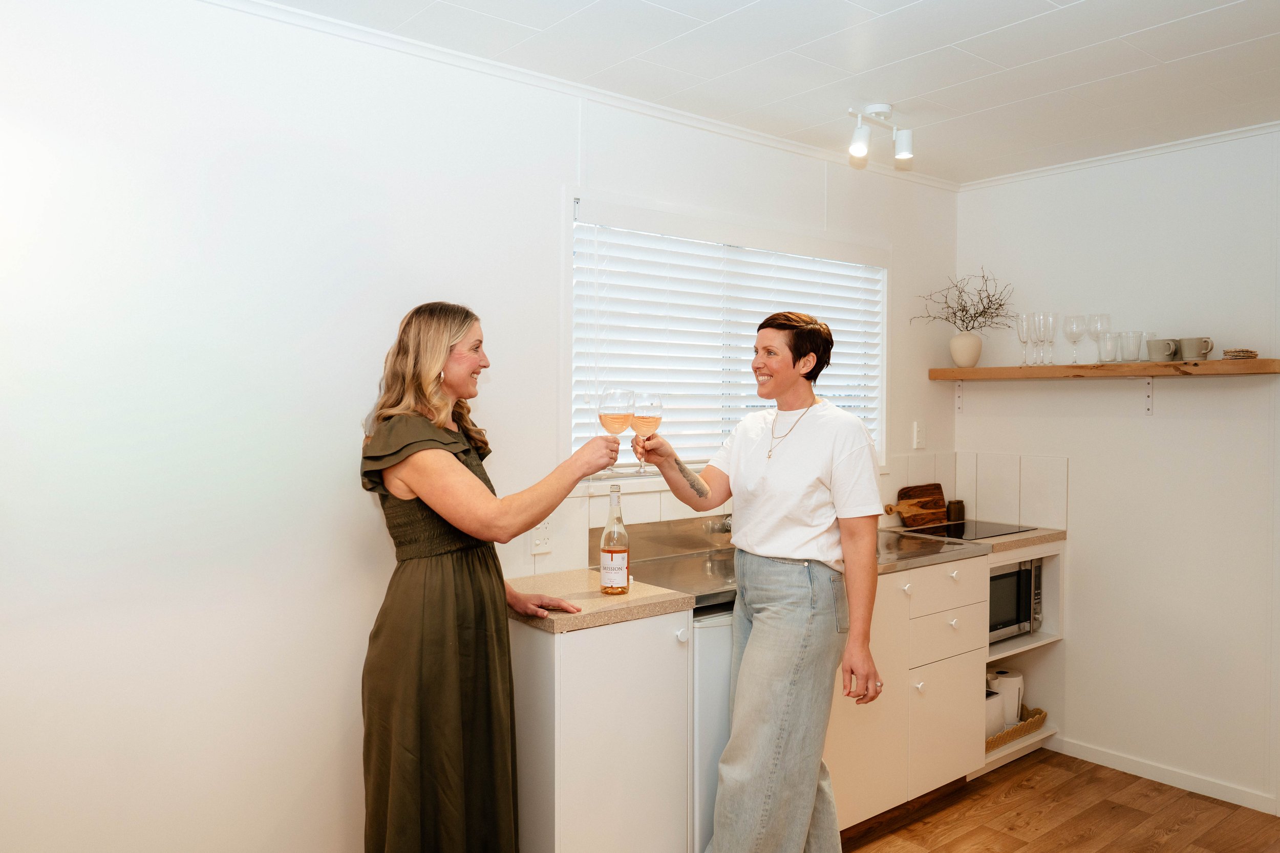 Two women in a kitchen raising glasses of rosé wine in a toast, smiling at each other.