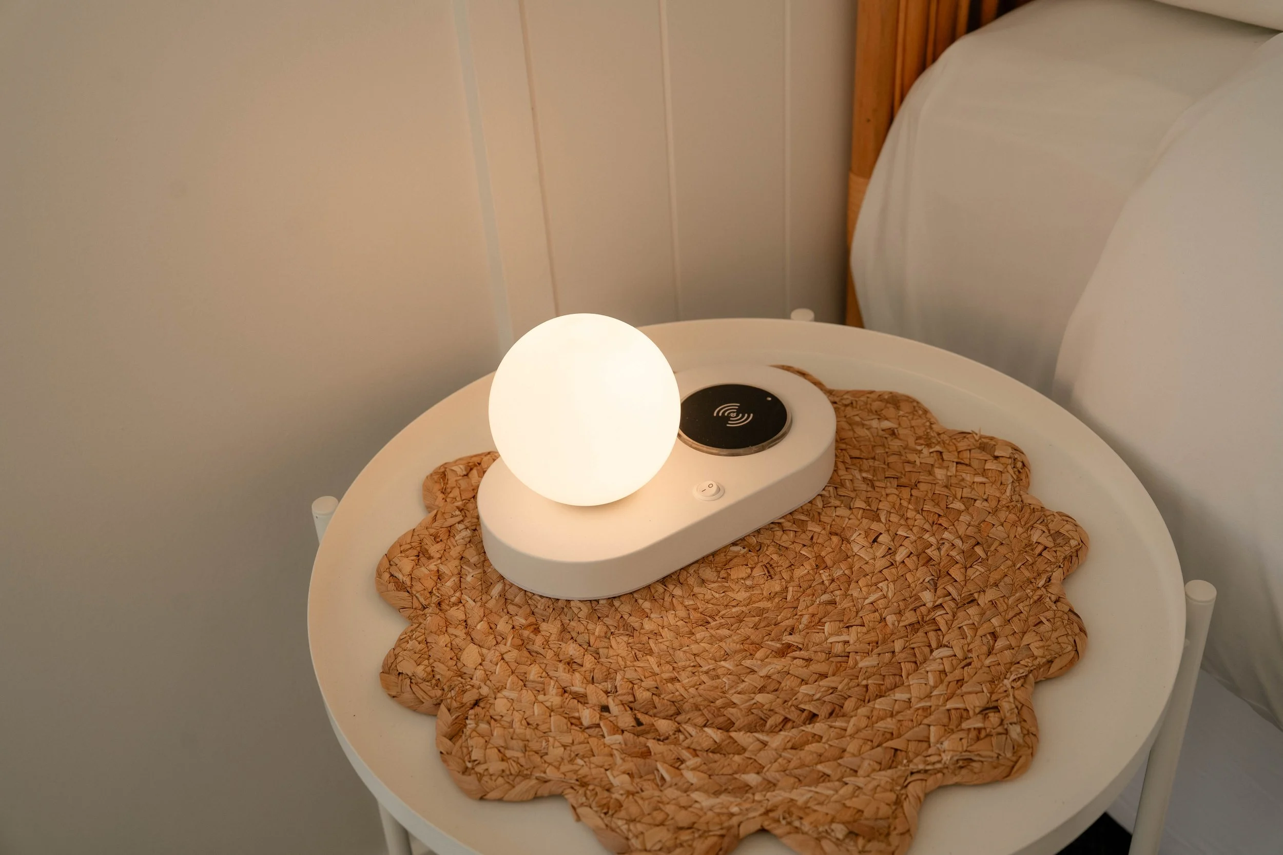 A small round white table with a woven placemat on top, a white spherical lamp, and a wireless charging pad. The table is next to a bed with a wooden headboard and a white bedsheet.