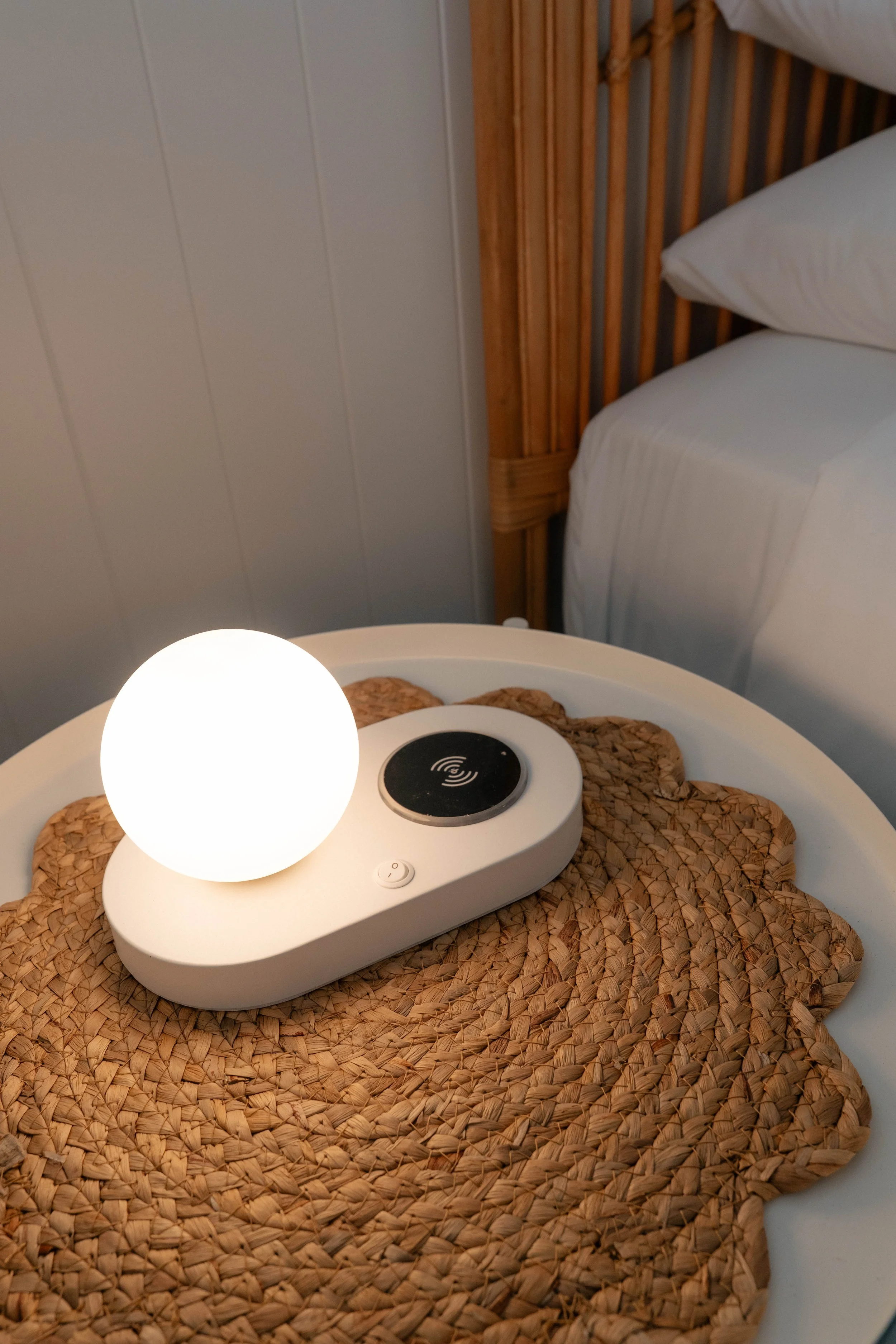 A round woven placemat on a white bedside table with a white bedside lamp and a wireless charging device, next to a bed with white linens and a wooden headboard.
